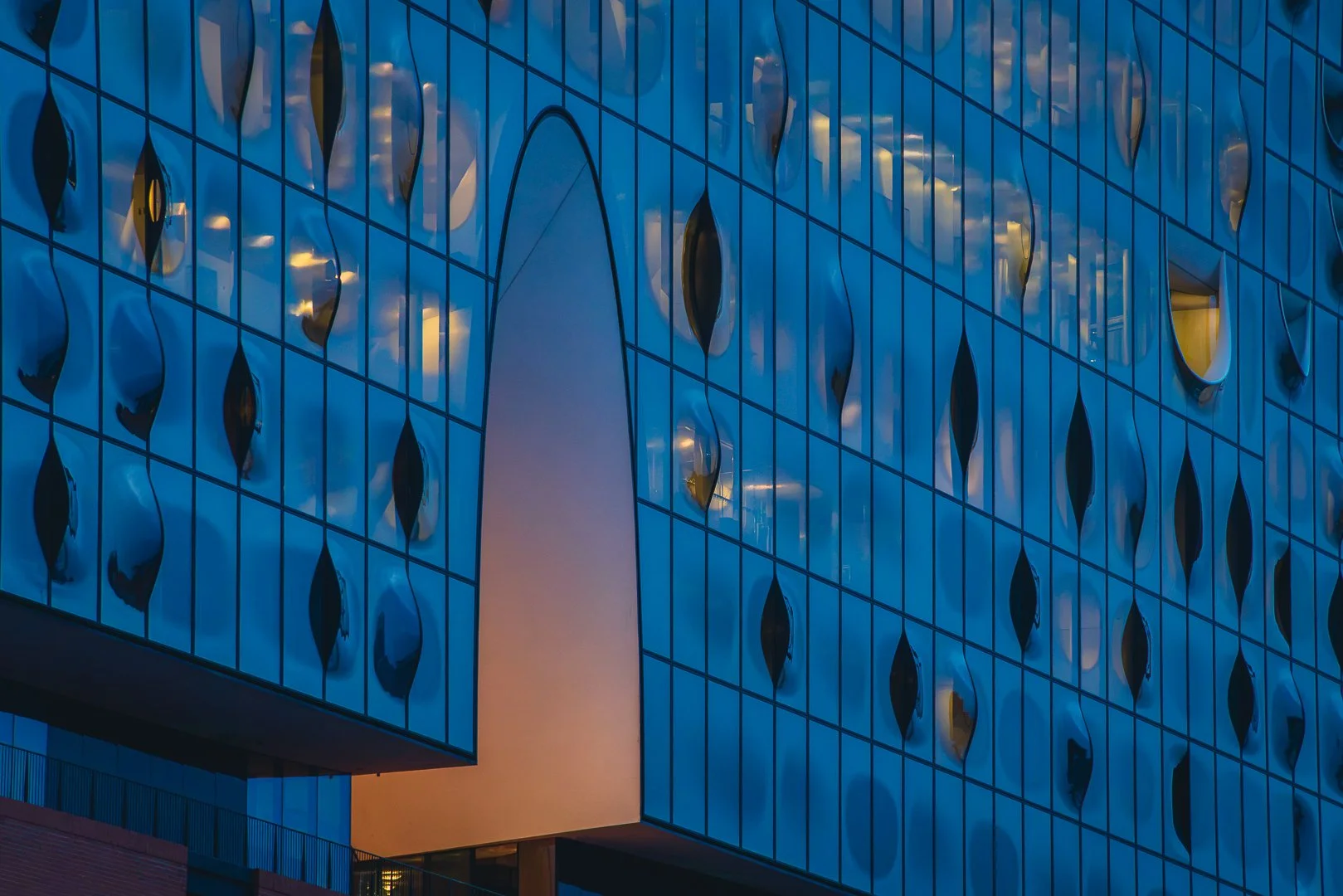 Architectural detail of the Elbphilharmonie in Hamburg showing curved glass surfaces, dark windows and warm interior lights at blue hour.
