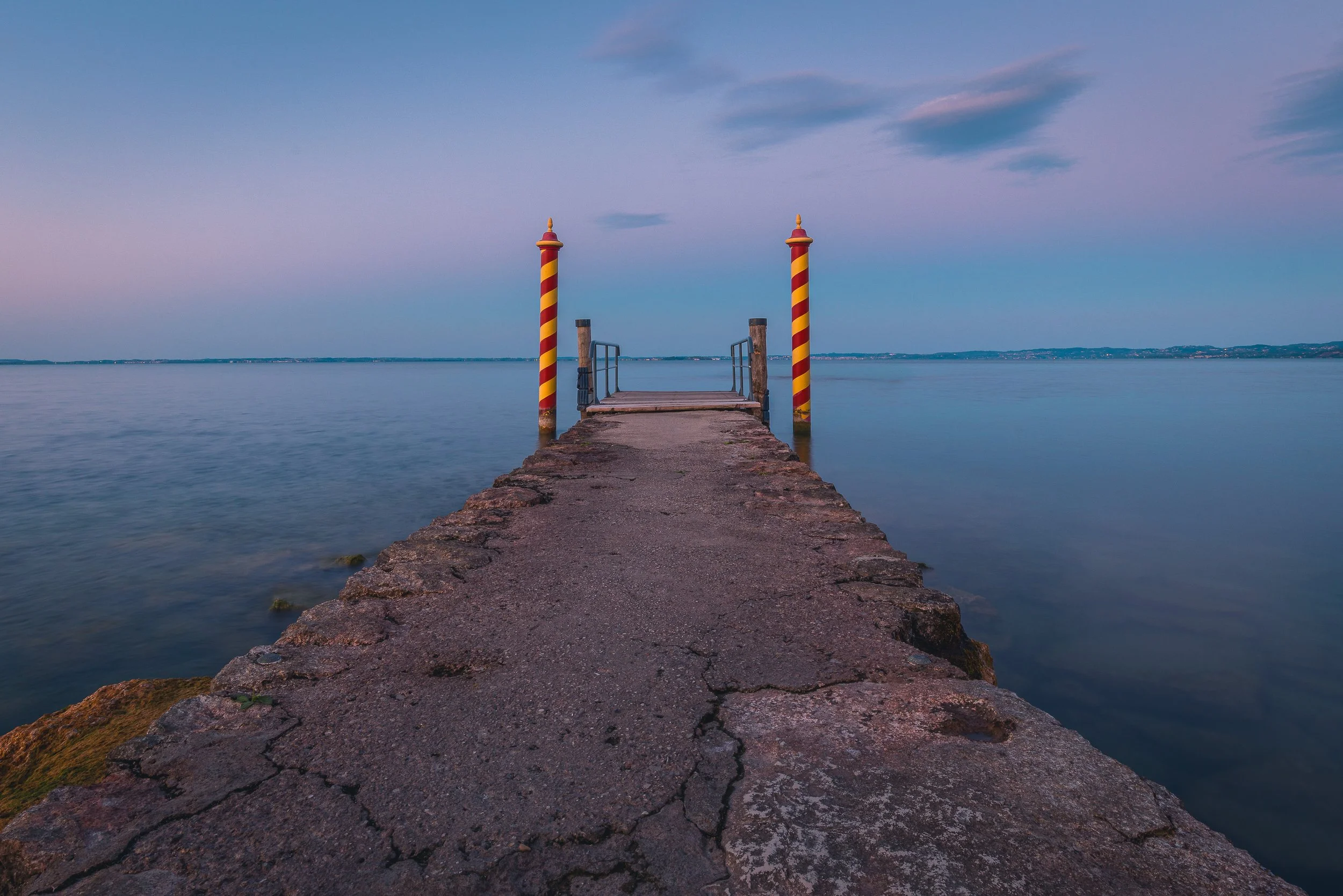 Stone jetty with striped posts extending into calm Lake Garda at blue hour