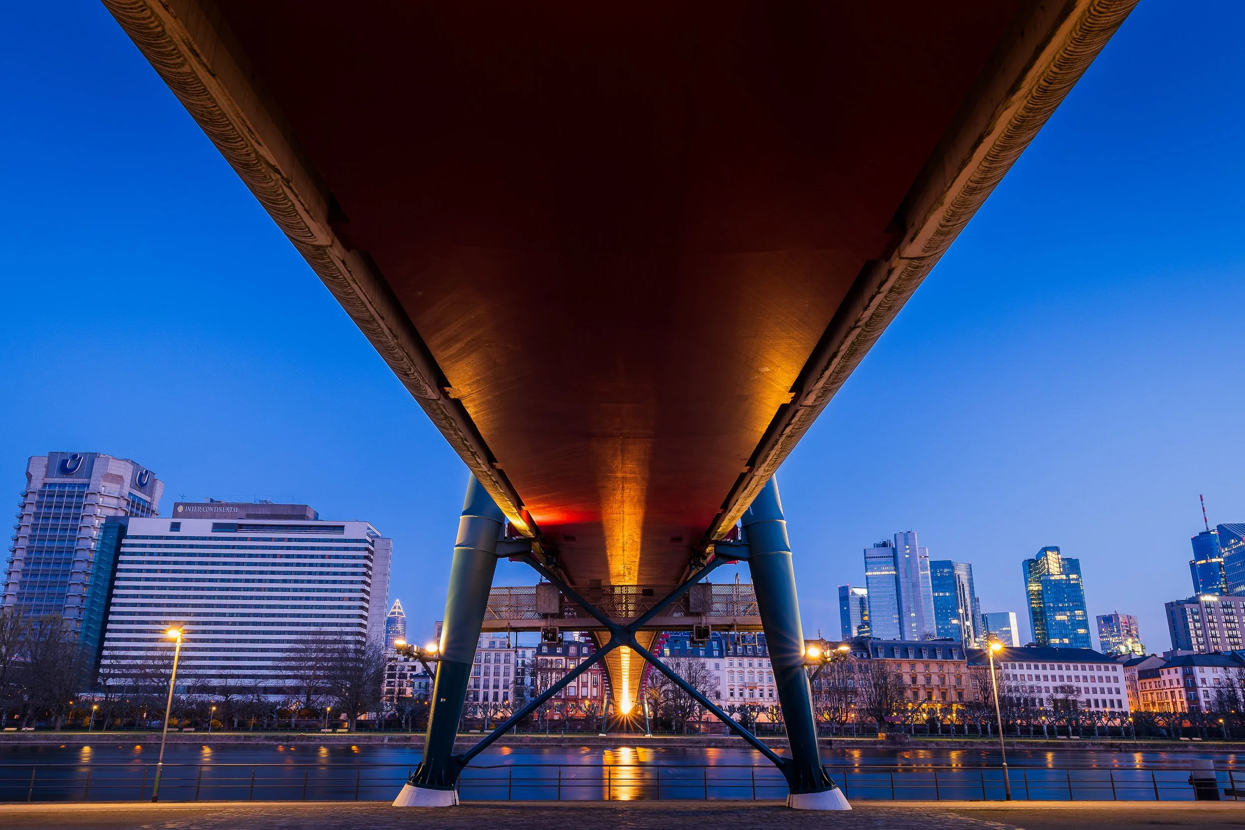 View beneath Holbeinsteg toward the illuminated Frankfurt skyline at blue hour