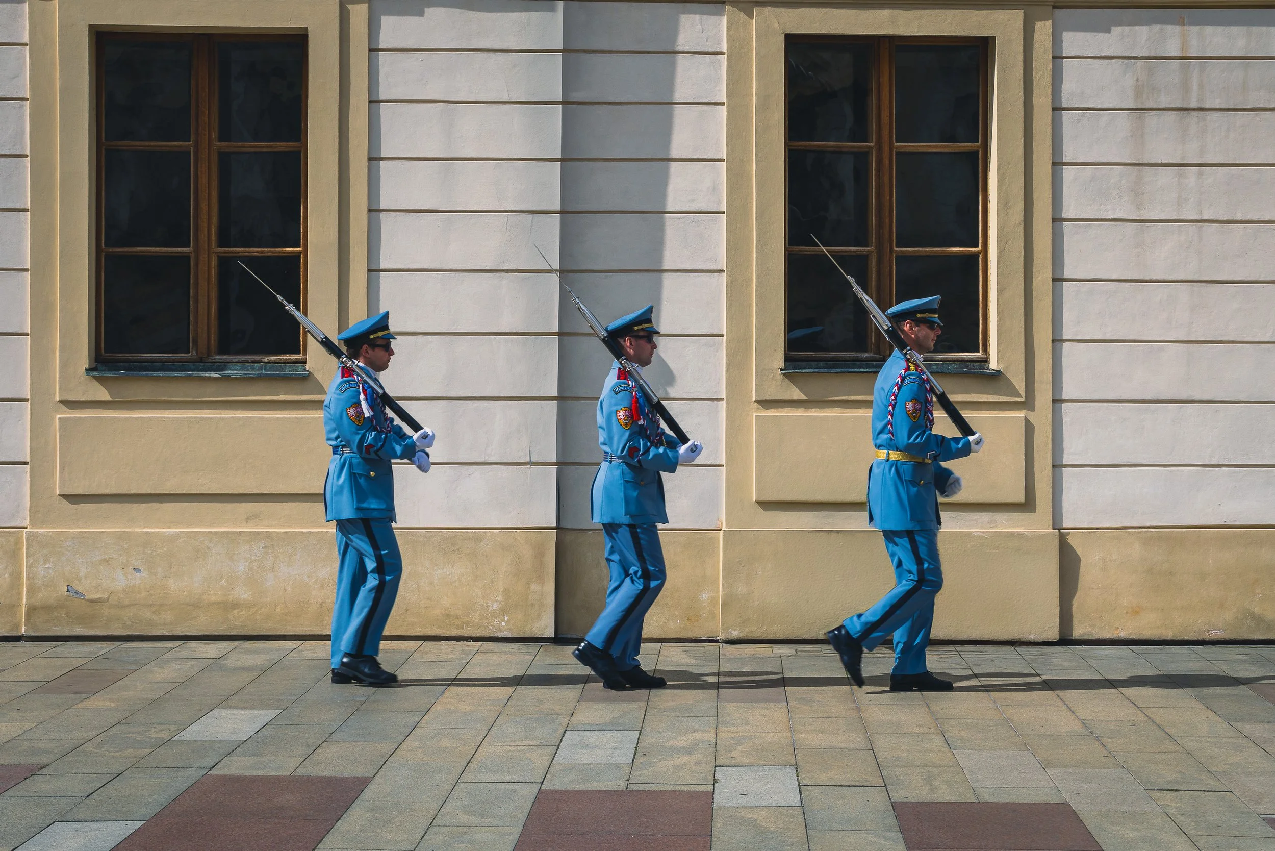Three ceremonial guards in blue uniforms marching past a pale stone facade in Prague