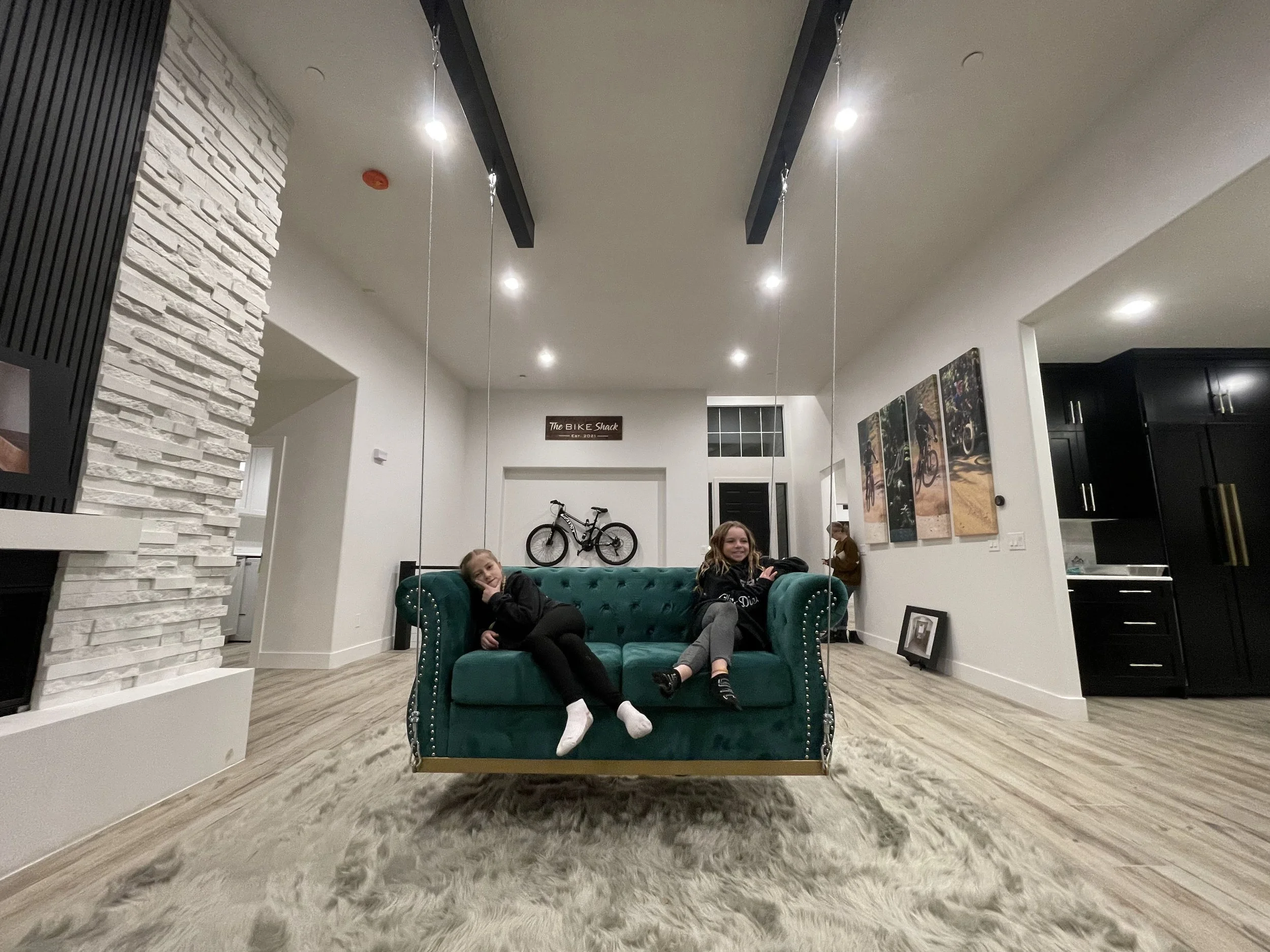Two girls are sitting on a teal tufted couch in a modern living room, with one girl lying down and the other sitting upright, smiling. The room has a white textured wall, black ceiling beams with hanging lights, and a mounted bicycle is visible in th