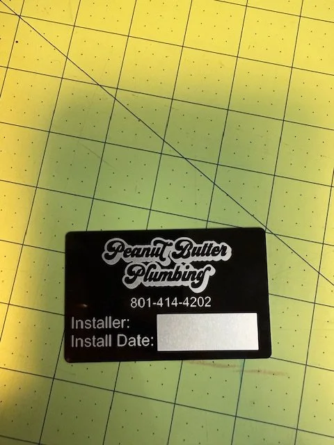 A black sticker label with white text reading 'Peanut Butter Plumbing' and a phone number. Spaces for 'Installer' and 'Install Date' are blank. The label is placed on a yellow cutting mat with grid lines and measurement marks.