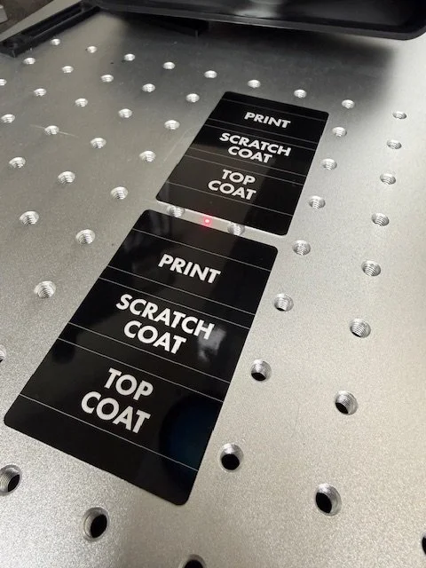 Black laser cut signs on a metallic surface with evenly spaced holes, indicating the printing of print, scratch coat, and top coat.