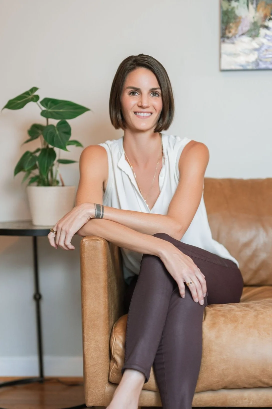 Leslie Roach, LCSW, wearing a white sleeveless blouse and dark pants, sitting on a tan leather sofa, smiling, with a green potted plant and a painting on the wall behind her.