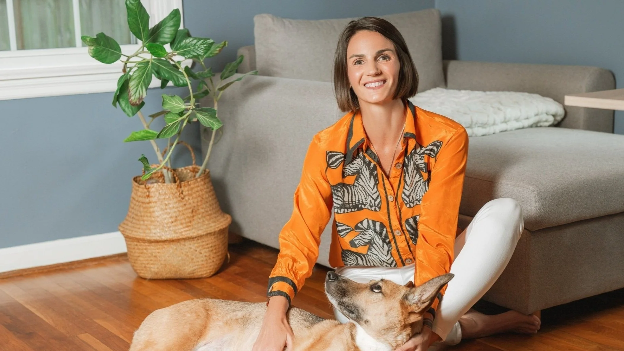 Leslie Roach, LCSW, sitting on the floor with a tan dog on a hardwood floor. She is wearing an orange shirt with a black and white leaf pattern. Behind her is a gray sofa, a green potted plant in a woven basket, and a window.