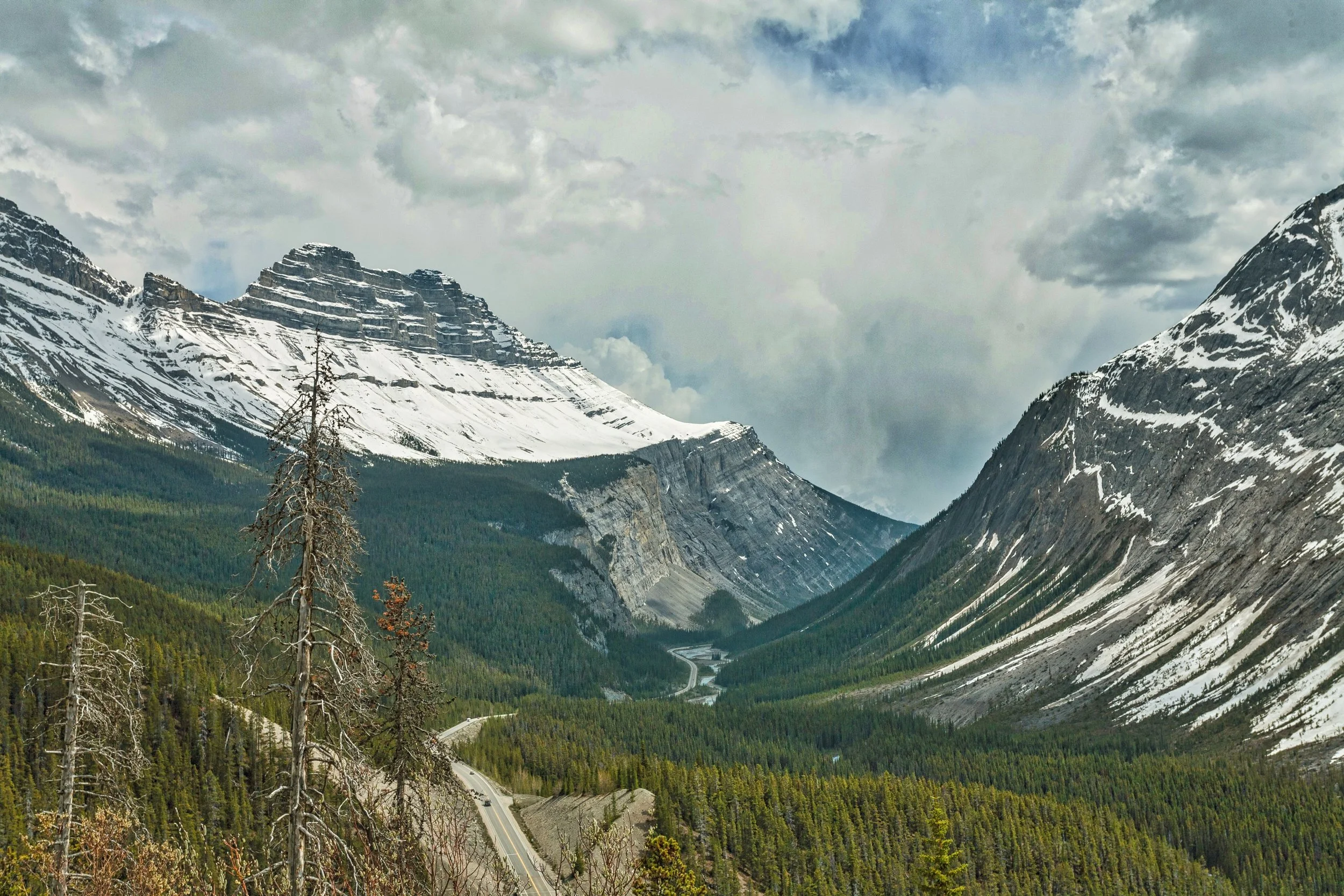 Snow-capped mountains surrounding a lush green valley with a winding river and a road, under a cloudy sky.