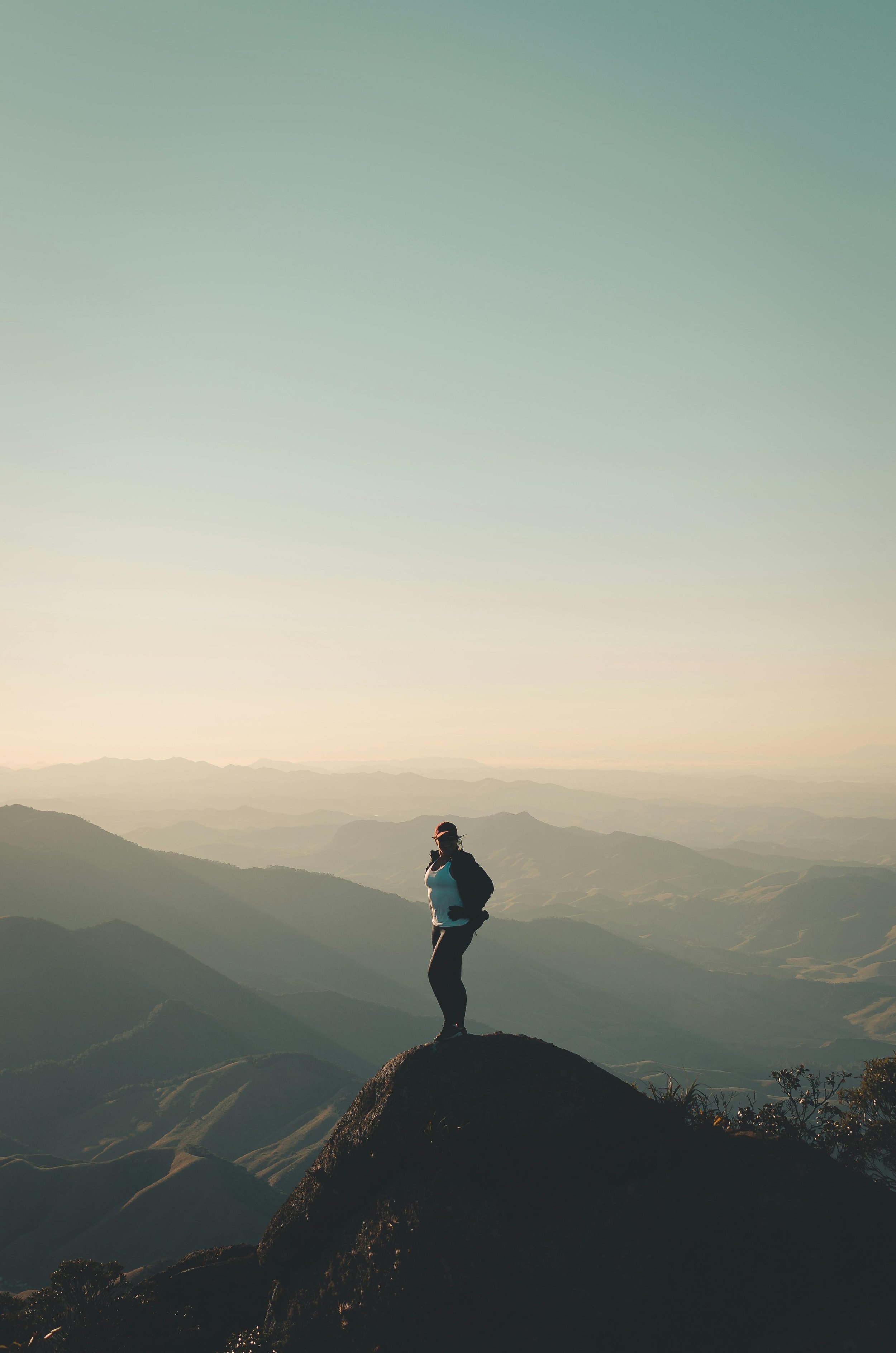 Person standing on a large rock at the top of a mountain, with a mountainous landscape and a pastel-colored sky in the background.