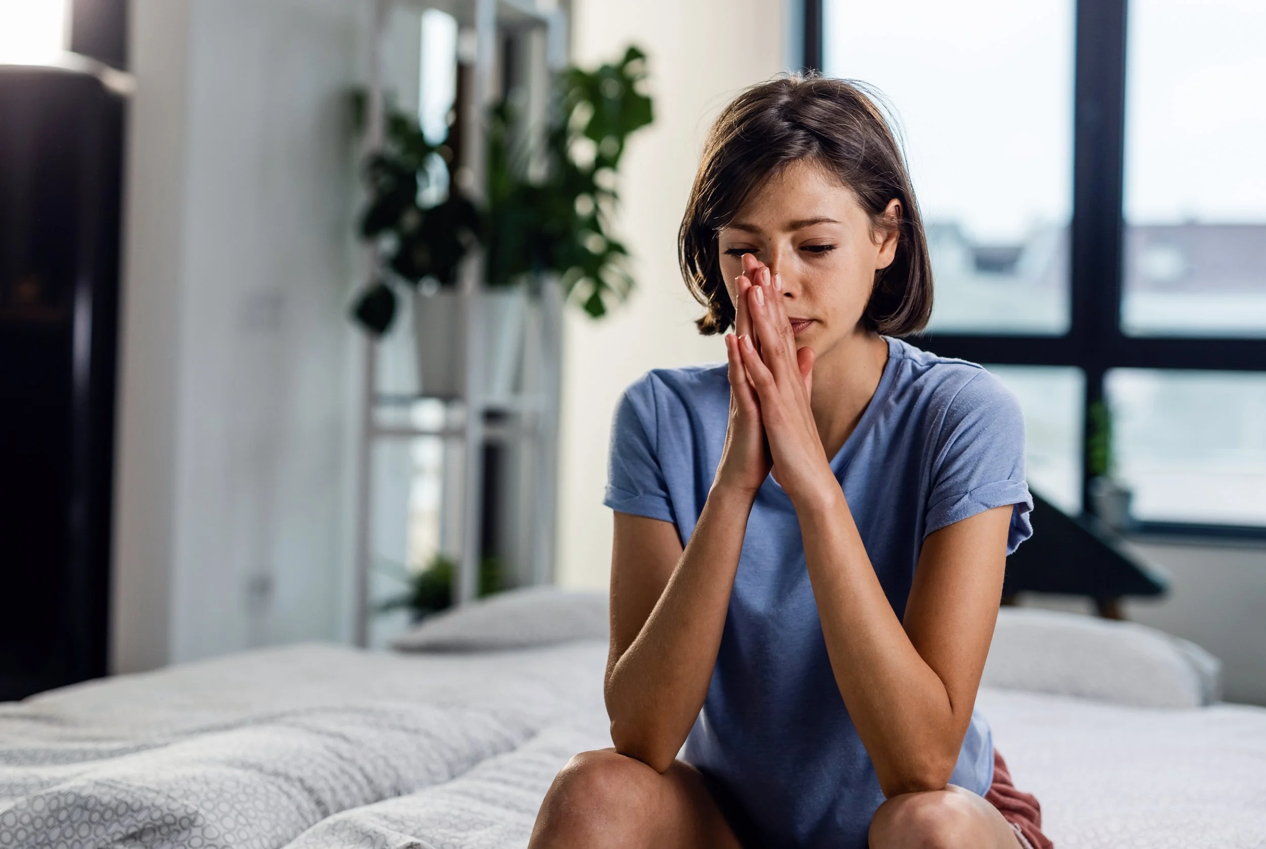 A young woman sitting on a bed in a modern room, looking distressed with her hands clasped near her face.