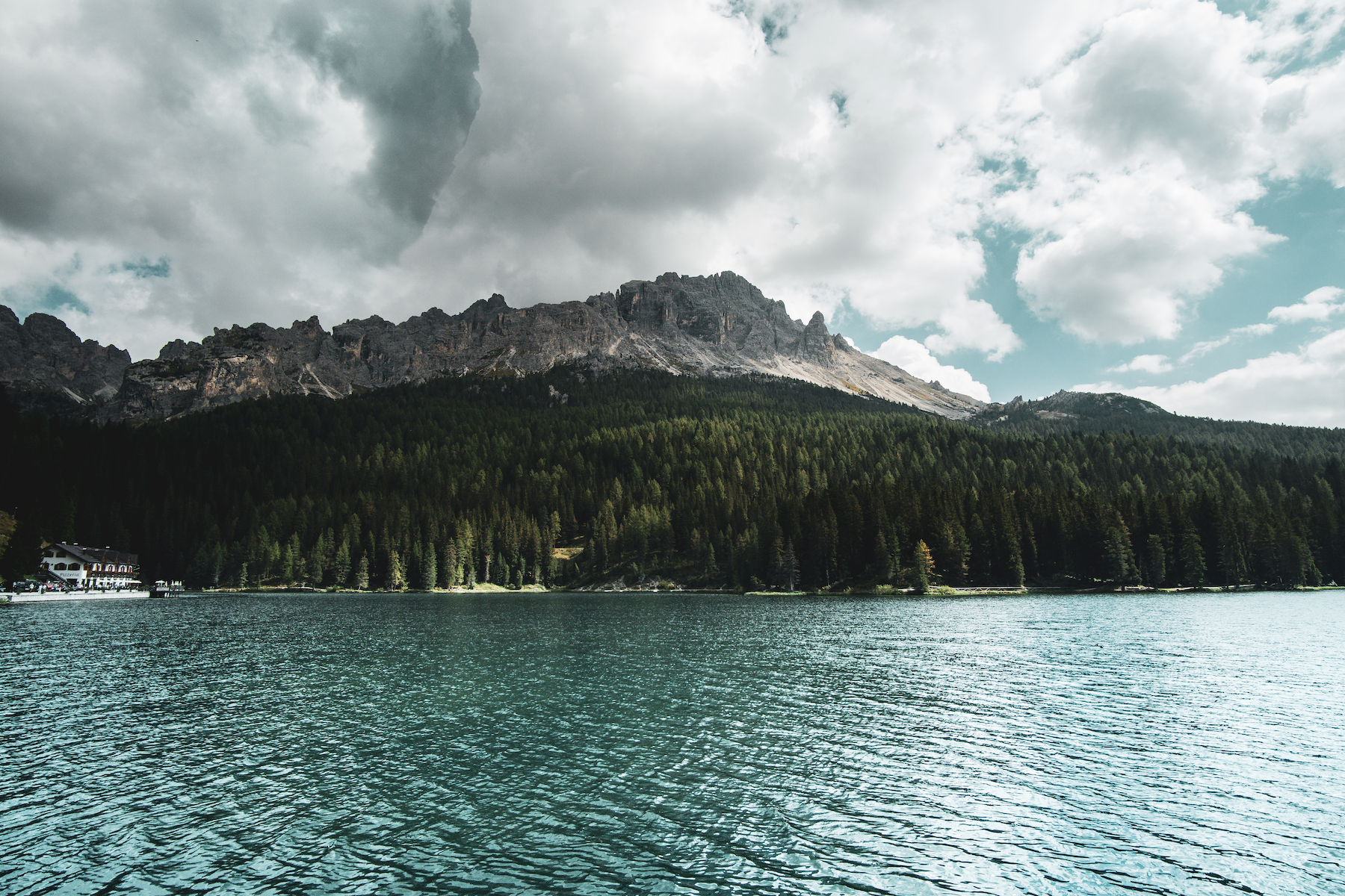 Scenic view of a mountain with clouds overhead, a dense forest on the slopes, and a lake in the foreground with rippling water.