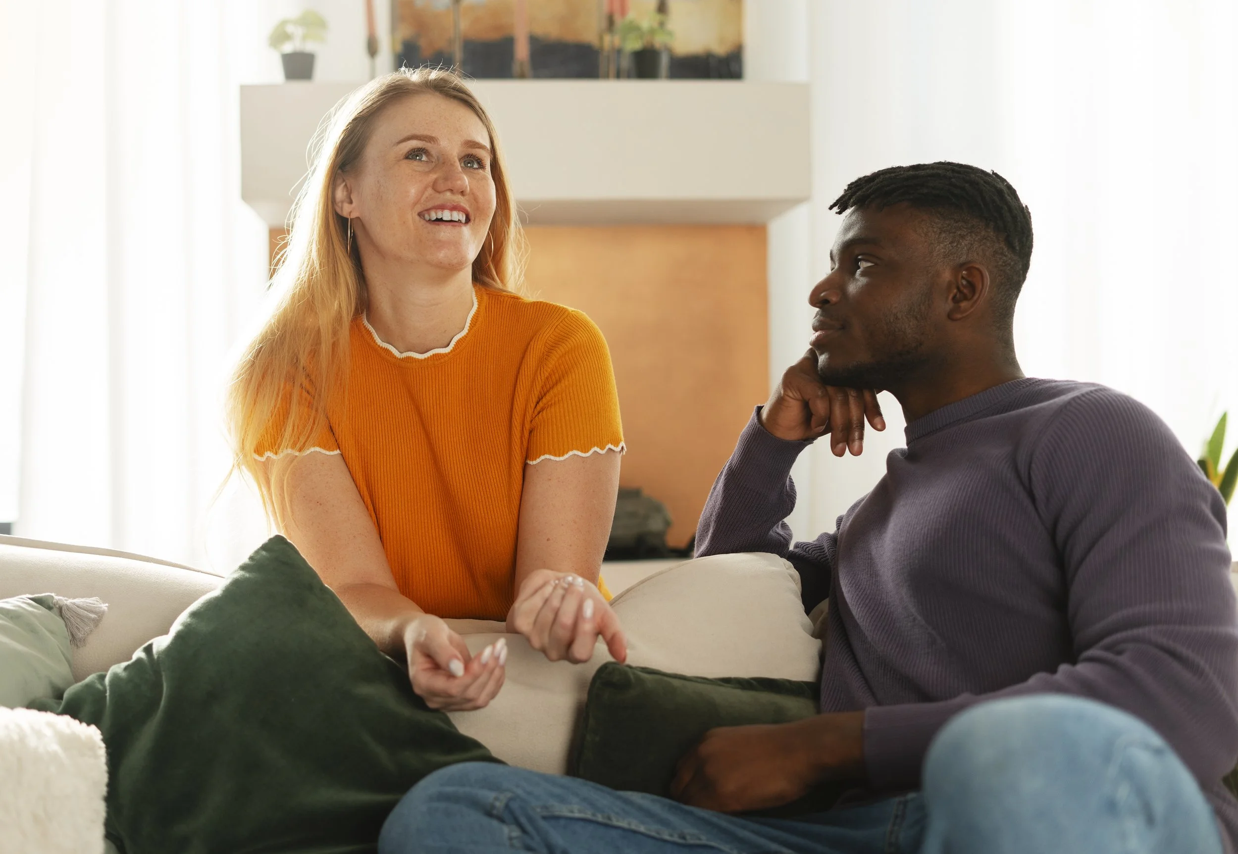 A woman with long red hair wearing an orange shirt smiling and talking to a man with short black hair and a beard, wearing a purple shirt, sitting on a sofa in a bright living room.