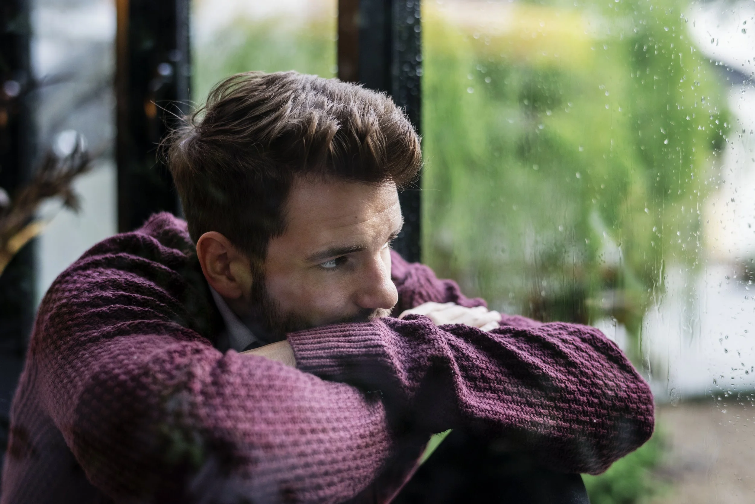 A man with brown hair and a beard resting his head on his crossed arms, looking out a rain-covered window, wearing a burgundy sweater.