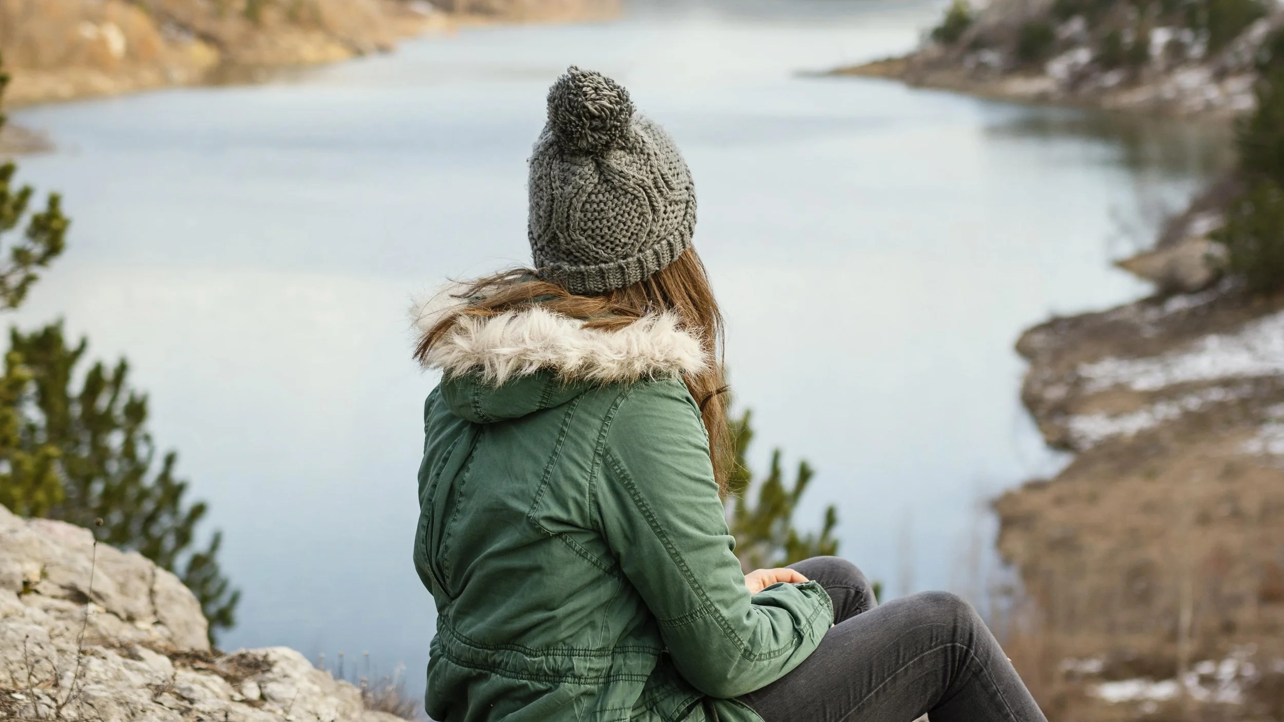 A woman in a green jacket and gray knit beanie sitting on a rocky ledge overlooking a calm river or lake, surrounded by sparse trees and a mountainous landscape.