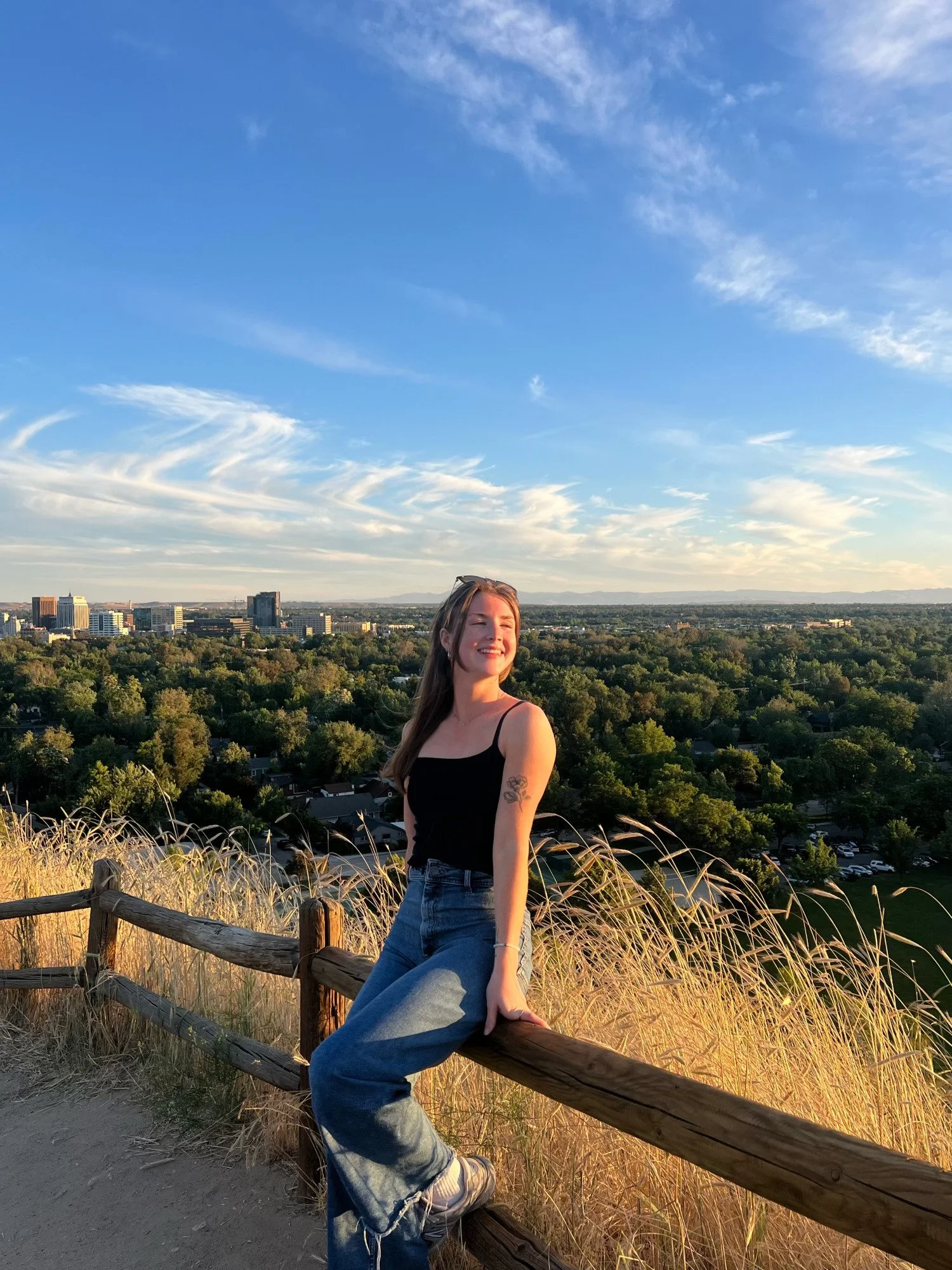 A young woman with long hair, wearing a black tank top and jeans, sitting on a wooden fence on a hill during sunset, overlooking a cityscape with tall buildings and a forested area.