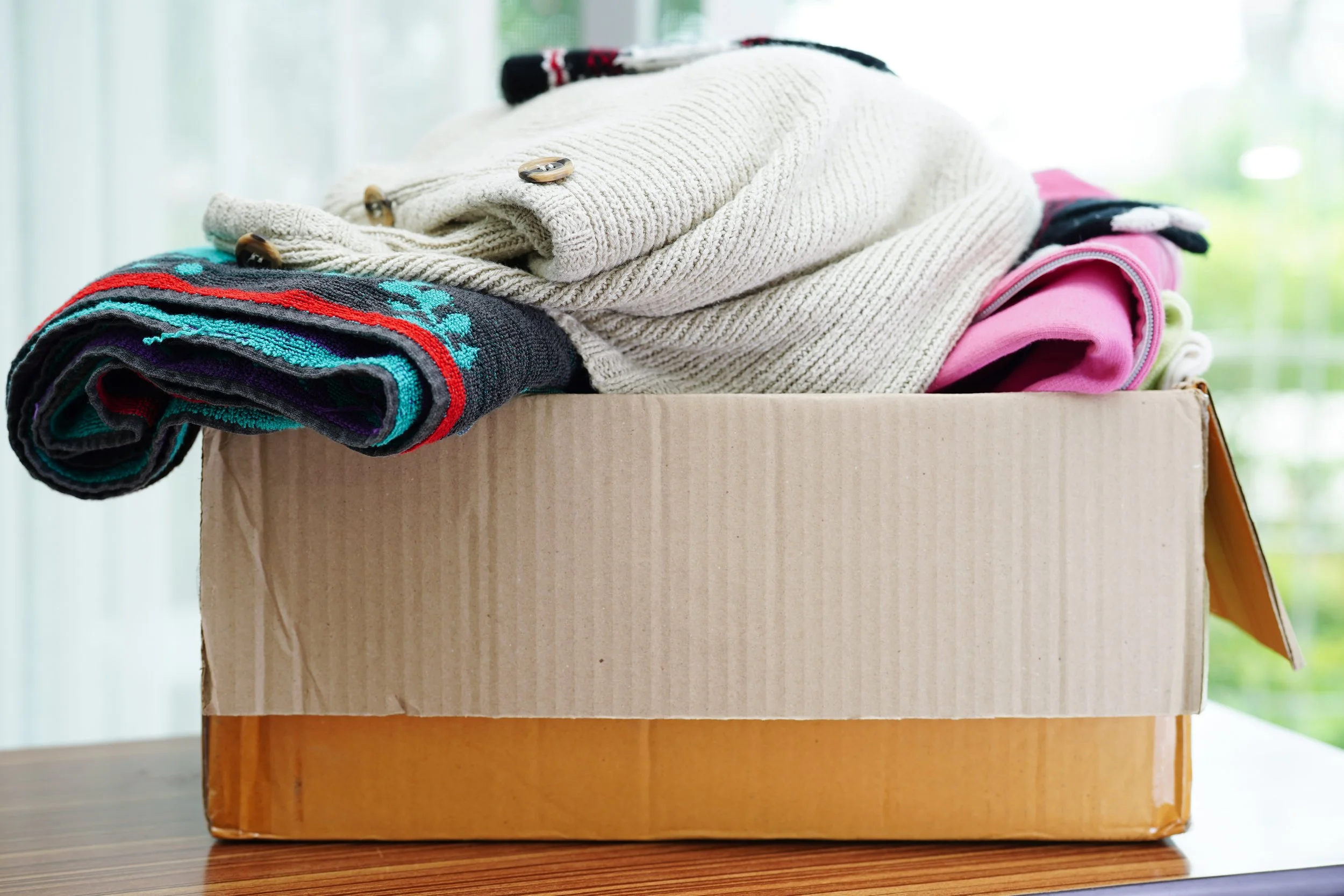 A cardboard box filled with folded clothes, including sweaters and socks, on a wooden surface with a blurred window in the background.