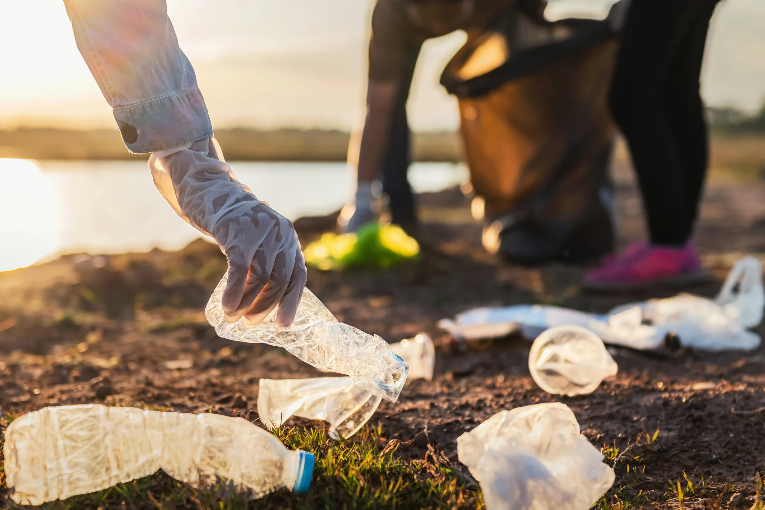 Person collecting plastic trash, such as water bottles and plastic debris, outdoors near a body of water during sunset.