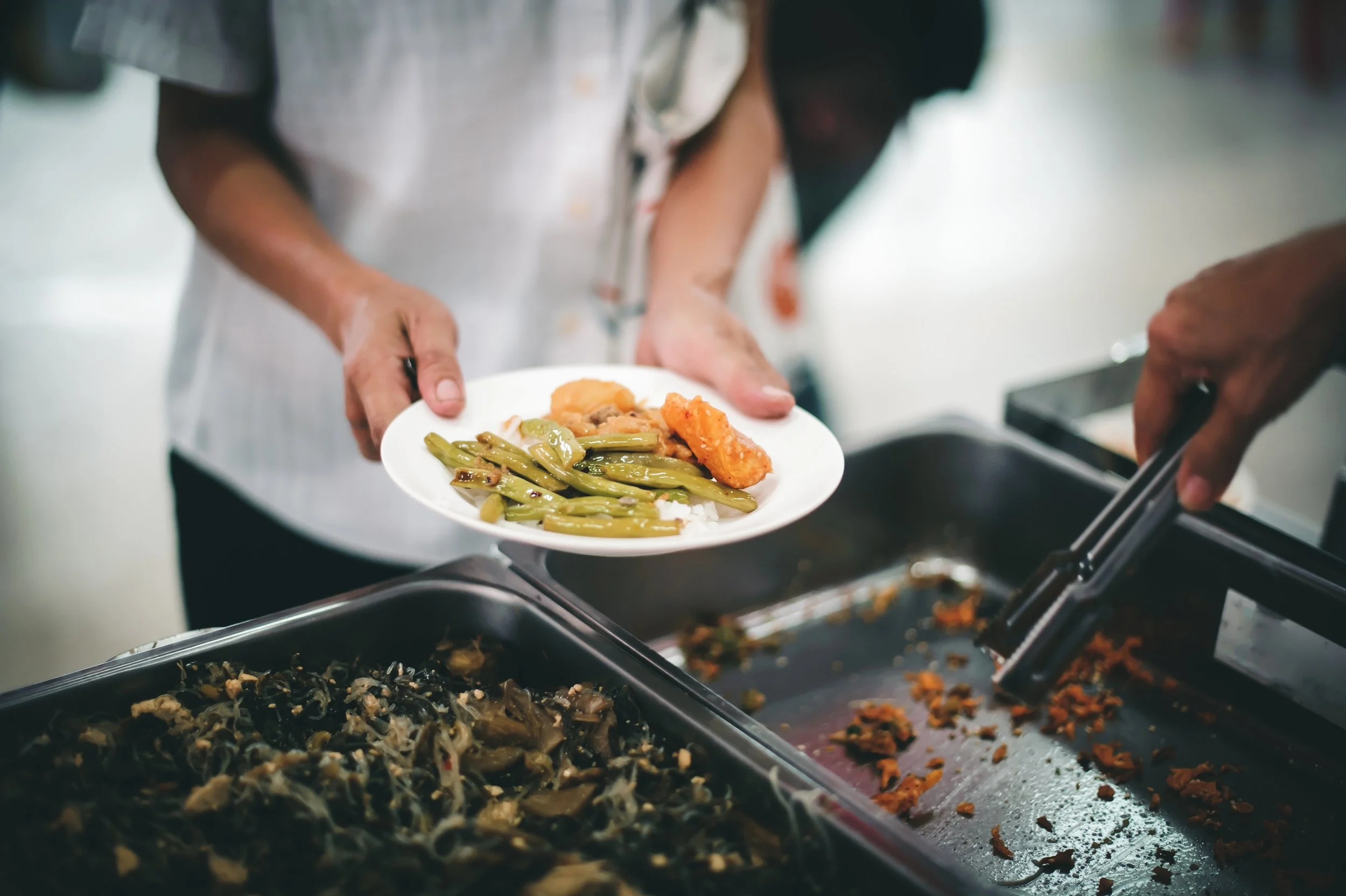 Person below receiving a plate of cooked green beans, fried chicken, and mashed potatoes from another person at a cafeteria or buffet-style food service station.