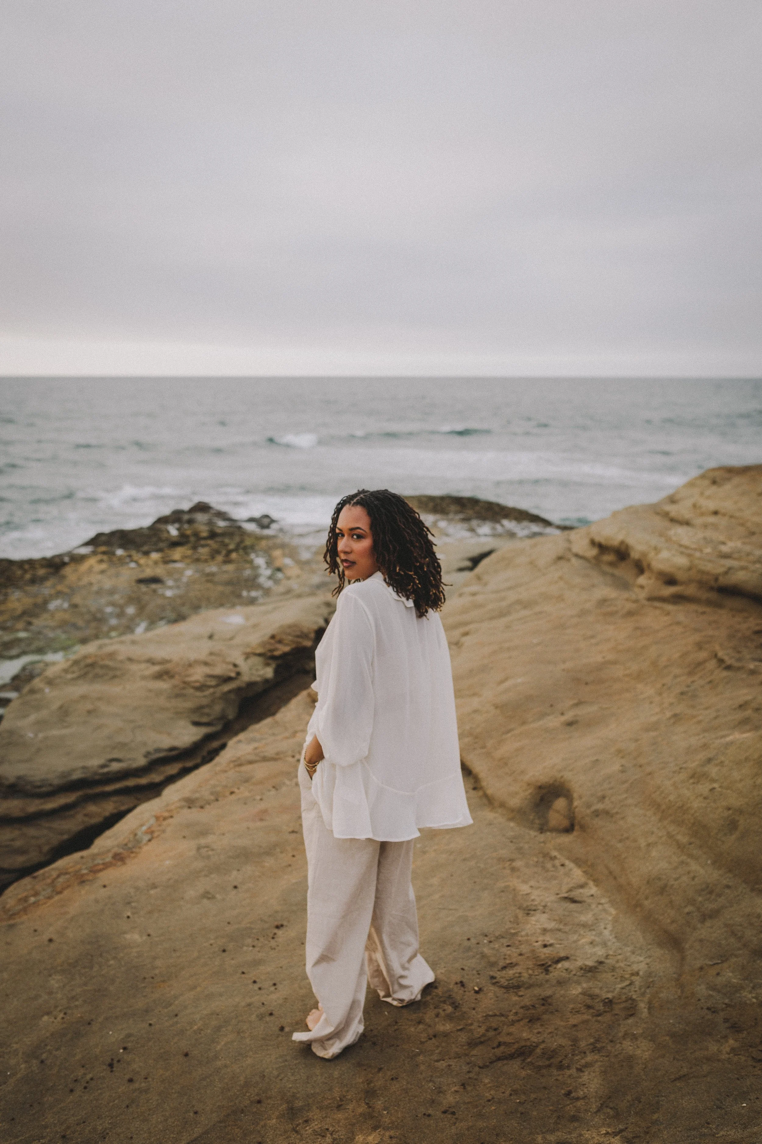 Woman with curly hair standing on rocky terrain at the beach, wearing a white loose blouse and beige pants, with the ocean and overcast sky in the background.