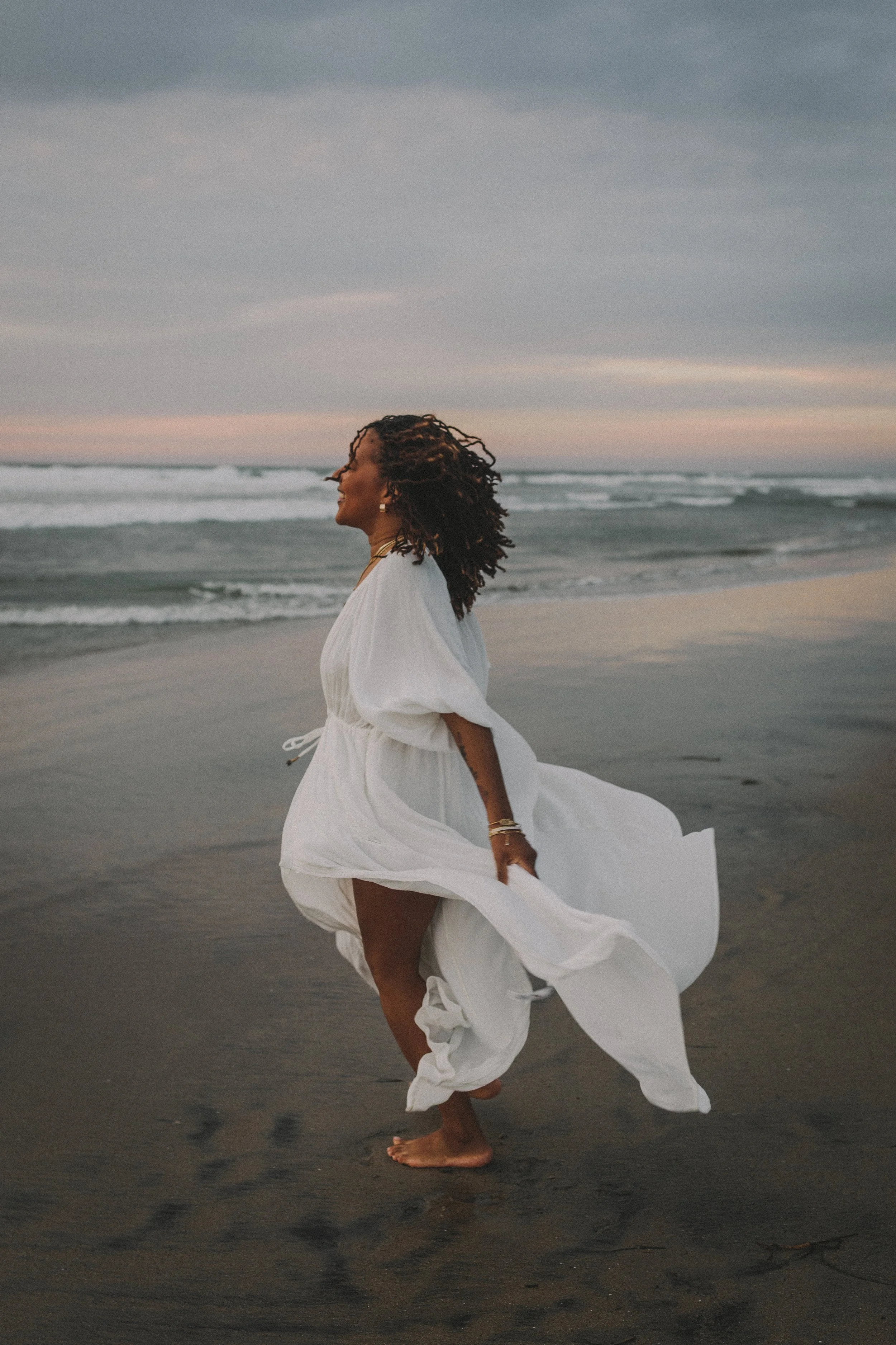 A woman in a white dress walking barefoot on the beach at sunset, with ocean waves in the background.