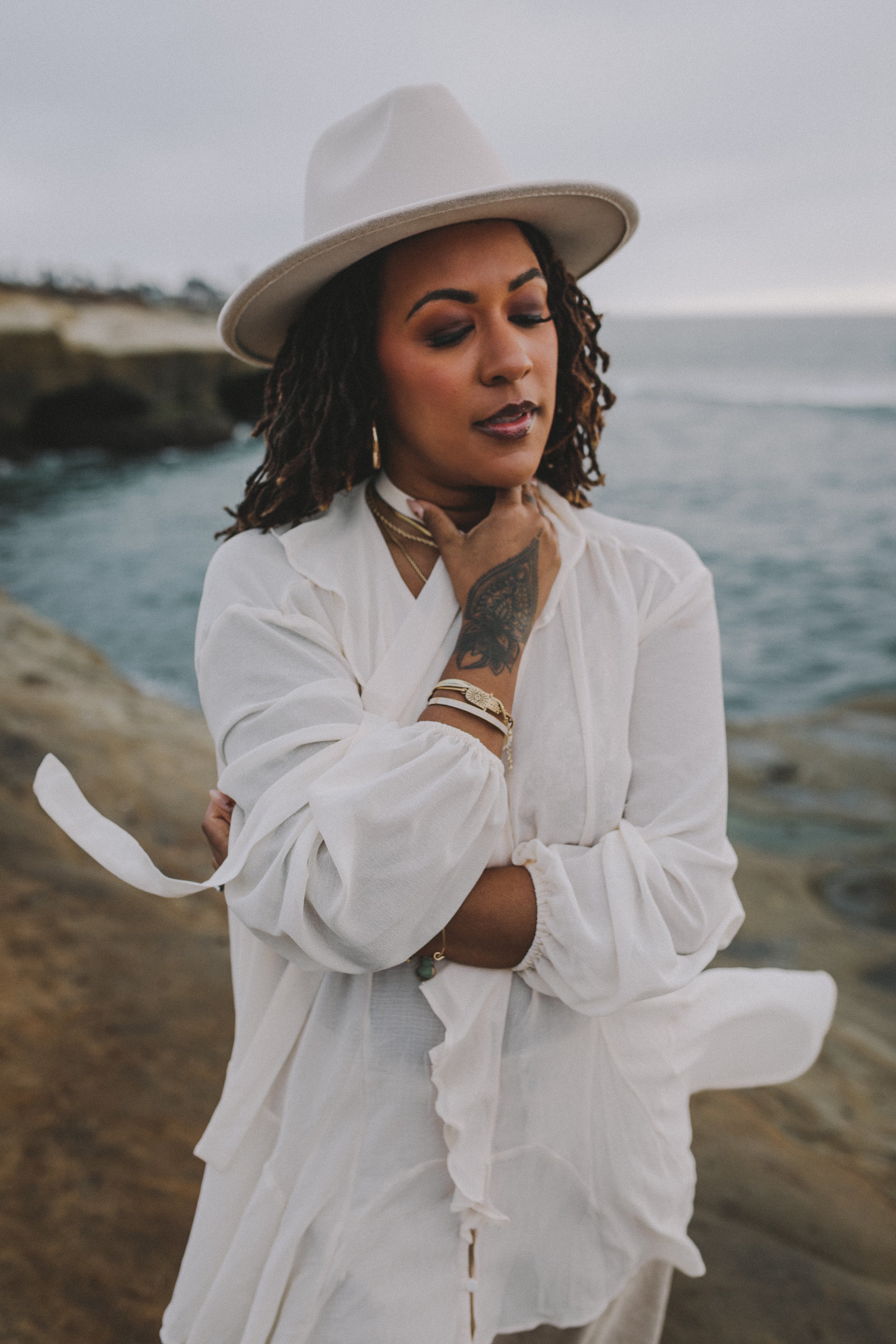 A woman with dreadlocks wearing a white hat and white clothing stands by the ocean, with her hand on her neck and her eyes closed.