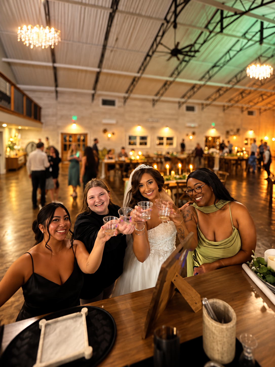 Four women, including a bride in a wedding dress and veil, at a wedding reception holding plastic cups in a rustic banquet hall with dim lighting and guests in the background.