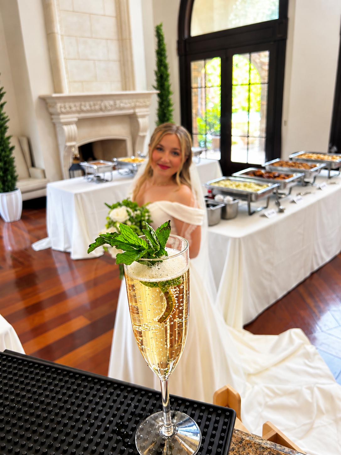 A glass of champagne garnished with mint and lemon slices in focus, with a smiling woman in a white dress and a buffet table with food in the background of an elegant room.
