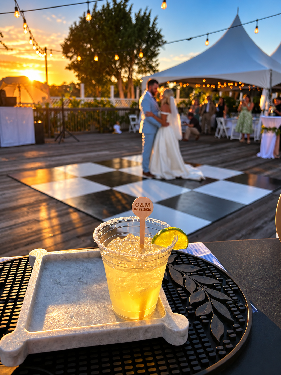 Wedding reception outdoors during sunset, with a couple dancing on a checkered floor and a Margarita drink with lime and a personal token on the table in the foreground.