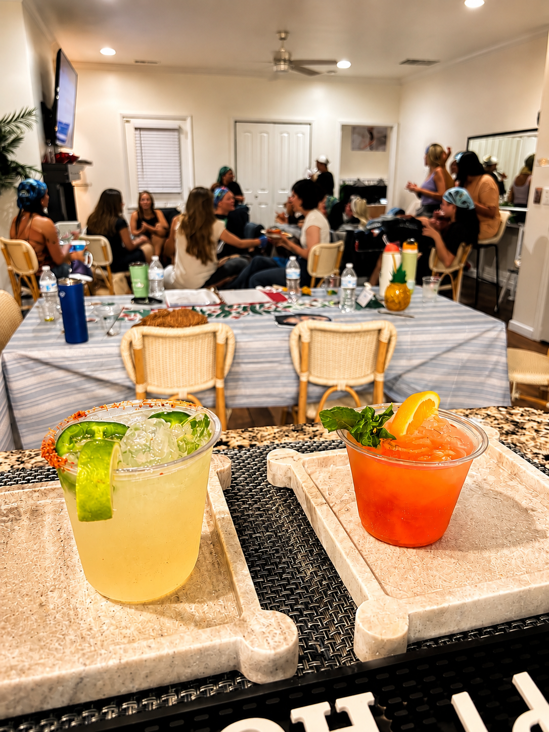 Two cocktails on trays in the foreground, with a group of people socializing in a room in the background.