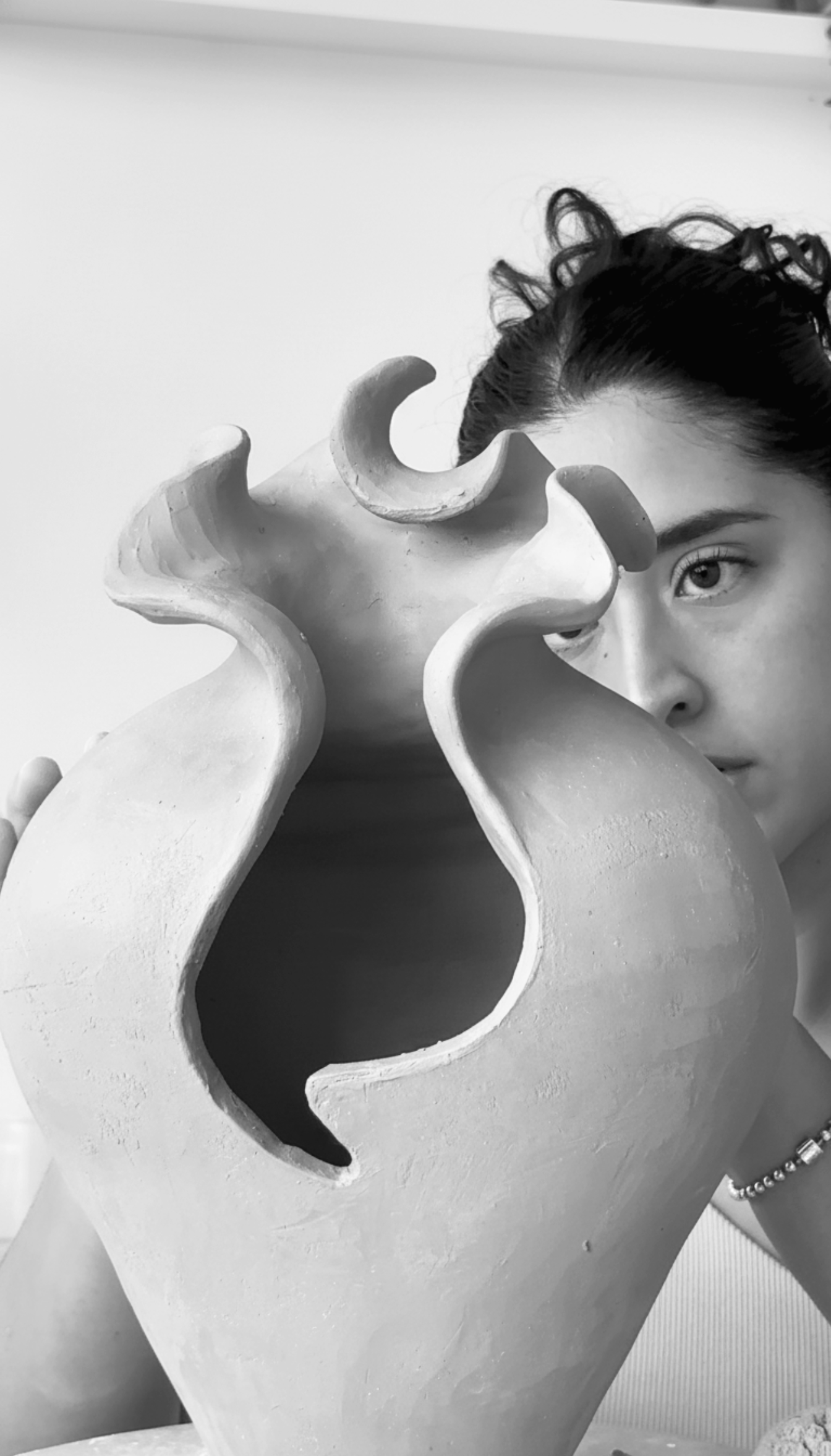 Black and white photo of a woman with dark hair hand building a large decorative ceramic vase with abstract, flowing cutouts, partially obscuring her face.