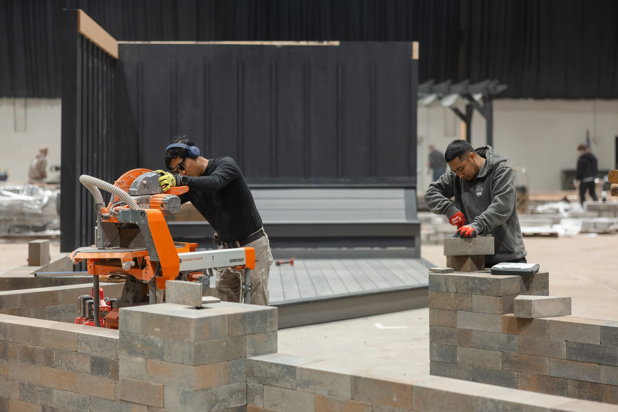 Two workers building a brick wall inside a construction site, one operating a brick cutting machine and the other stacking bricks.