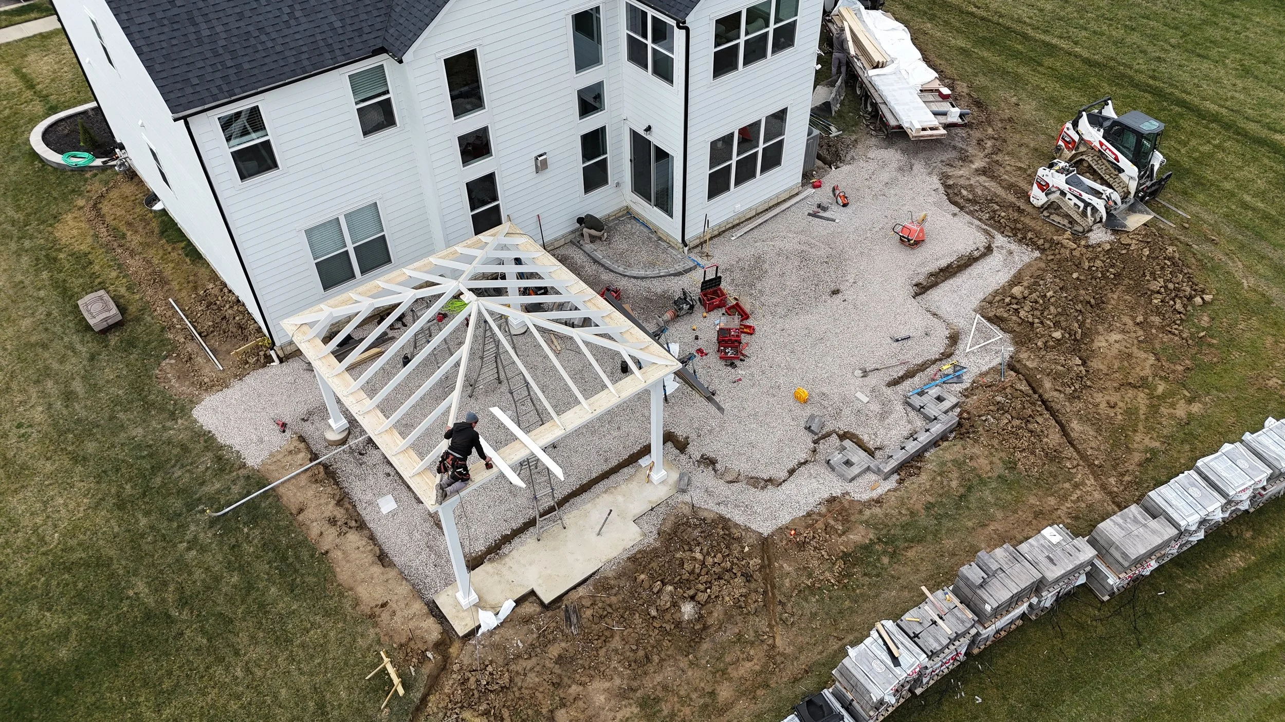 Aerial view of house construction site showing workers assembling a covered patio with white posts, gravel ground, construction tools, and machinery around the house, with a stack of tiles on the right side.