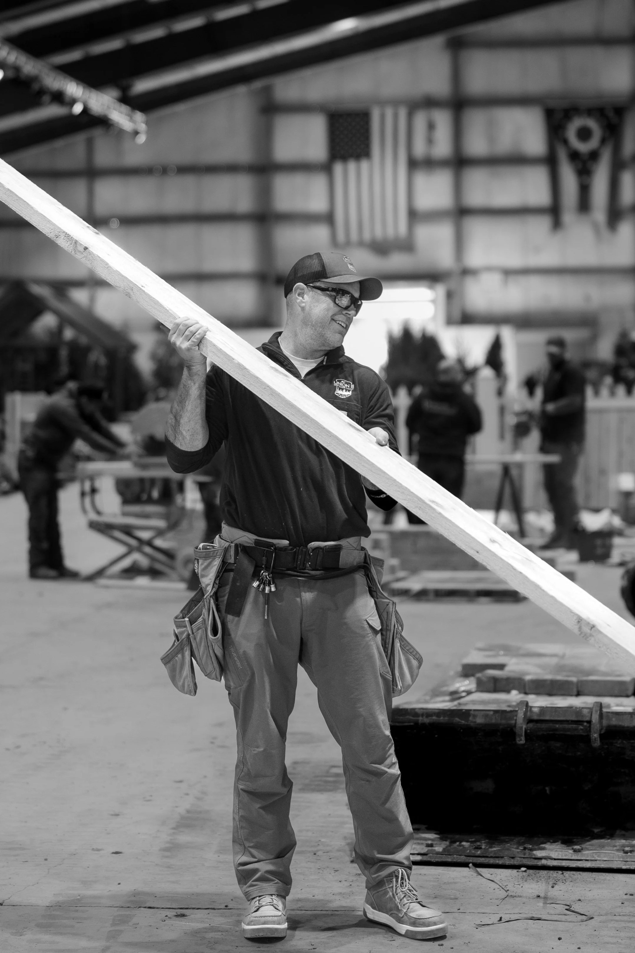 A man in a warehouse or workshop, wearing glasses, a cap, and a tool belt, carries a long wooden plank. In the background, there are several people working with tools, American flags, and banners hanging on the wall.