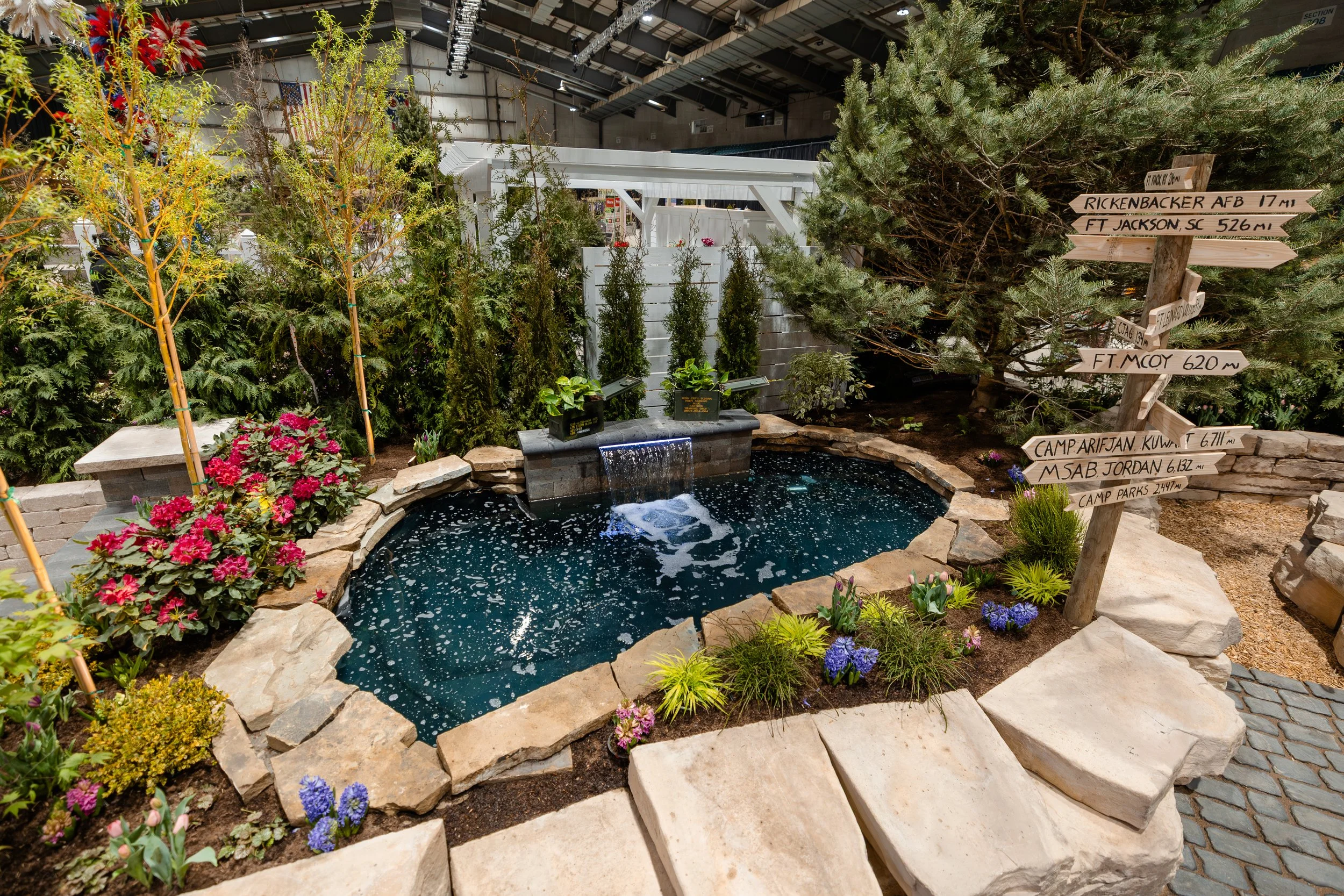 An indoor garden display featuring a small pond with a waterfall, surrounded by flowering plants, small trees, and a wooden directional signpost with distances to various locations.