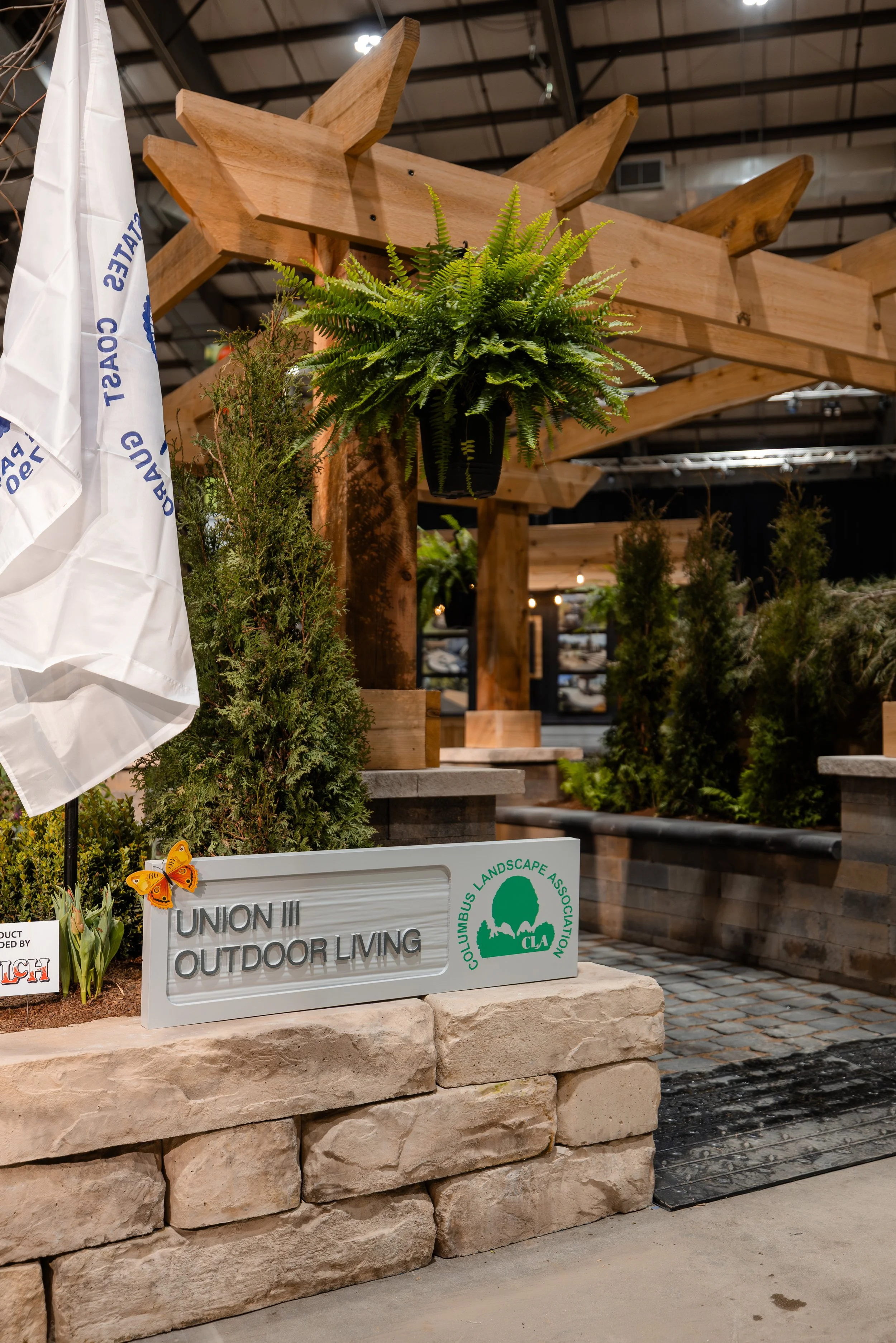 Indoor display with potted plants, a wooden structure, and a sign reading 'Union II Outdoor Living' by Columbus Landscape Association. The scene includes a hanging fern, small trees, and a white flag.