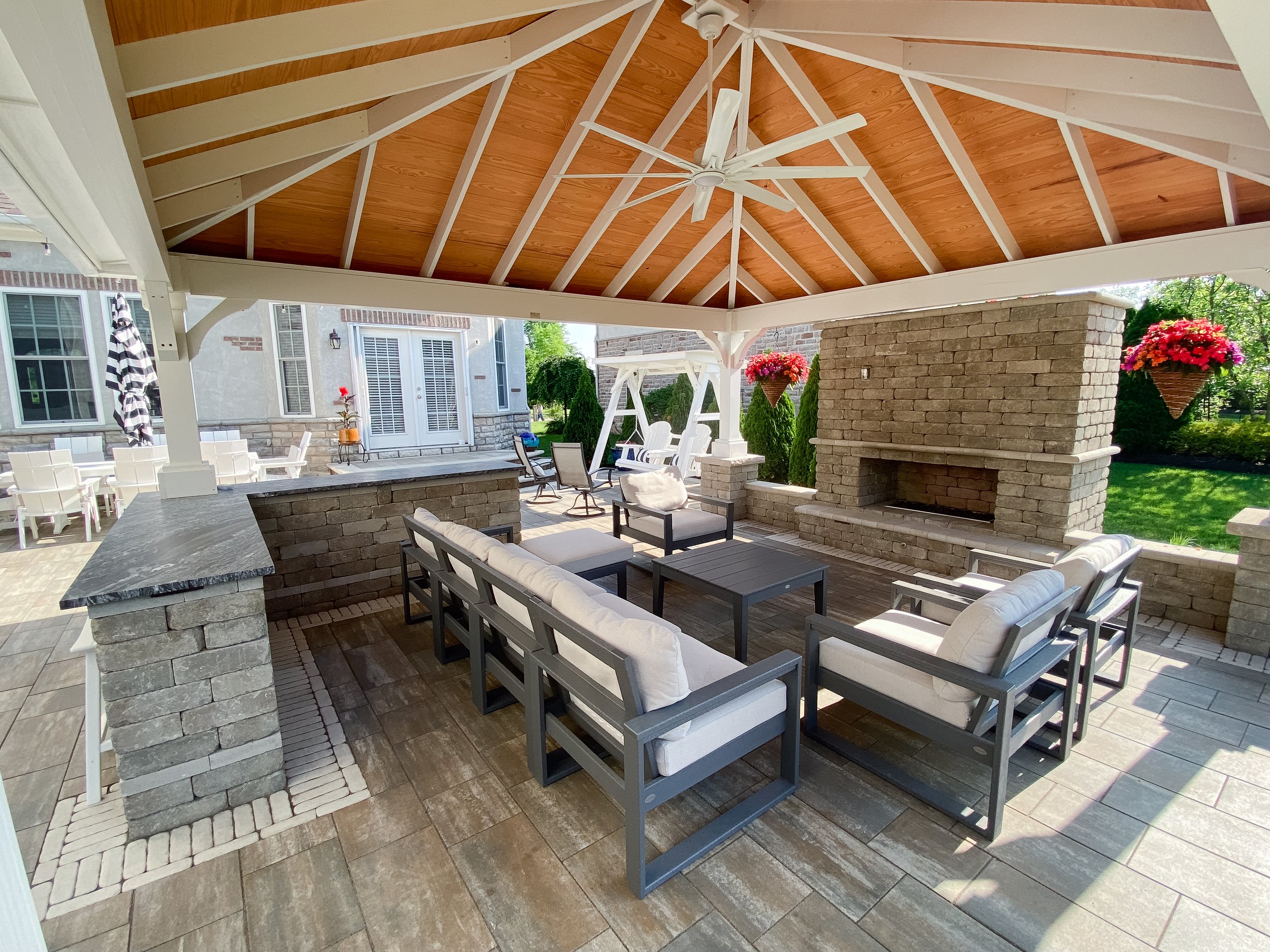 Covered outdoor patio with ceiling fan, brick fireplace, and outdoor seating including cushioned chairs and a coffee table, with hanging flower baskets and lush greenery in the background.