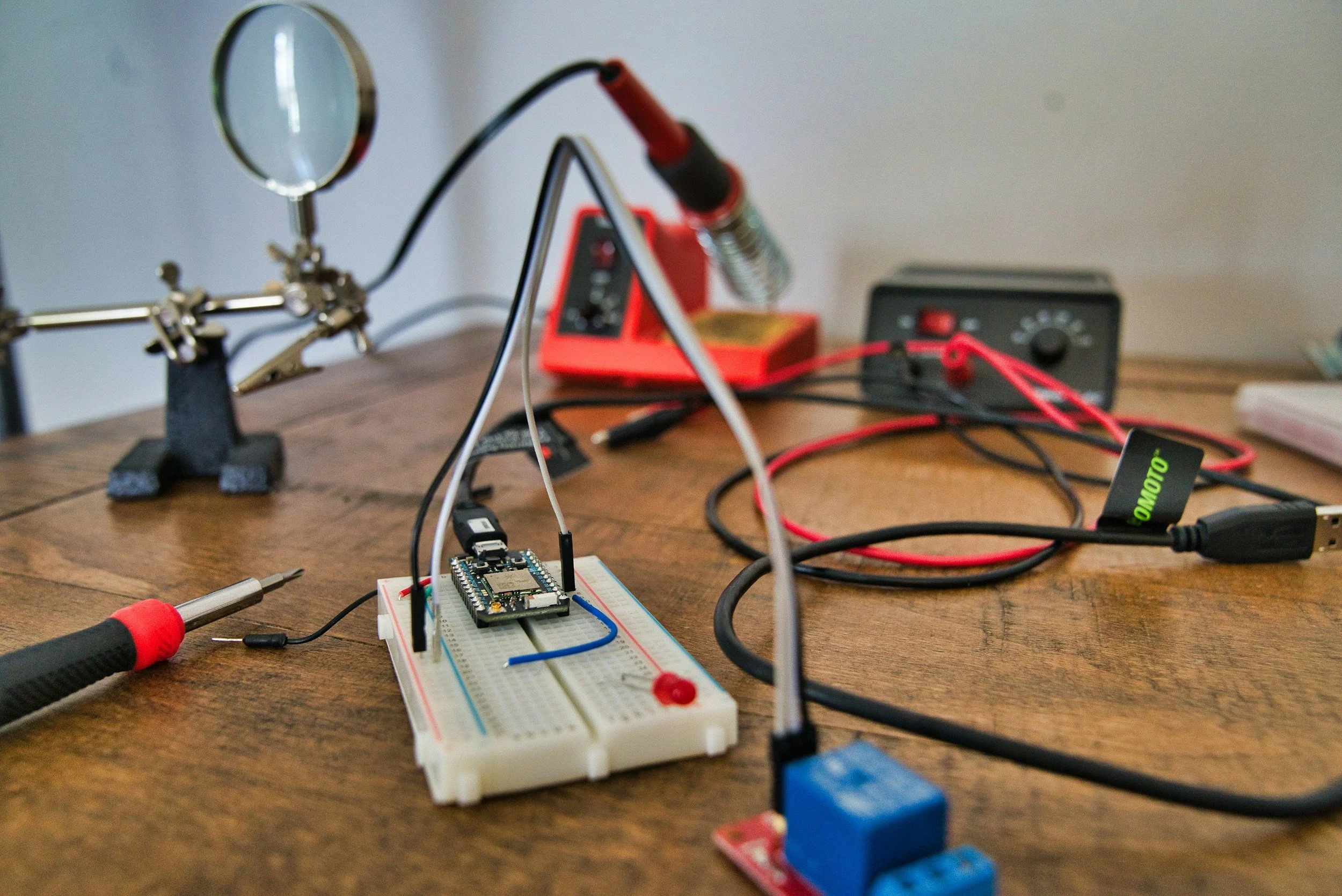 Electronics experiment setup on a wooden table including a breadboard with a microcontroller, wires, a soldering iron, a soldering station, and a magnifying glass with a stand.