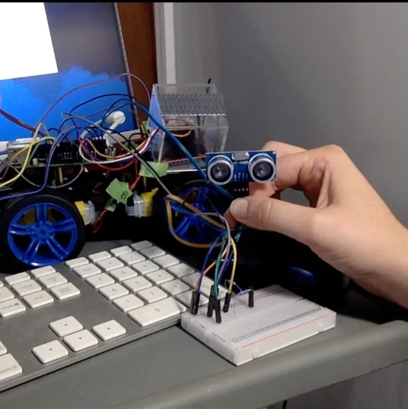 A person holding a small electronic sensor or component with a robot and various wires and electronic parts on a desk.