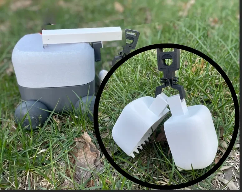 A children's toy snowplow with a gray body and black wheels, featuring a white scoop and two white salt or sand containers attached at the front, placed on grassy ground outdoors.
