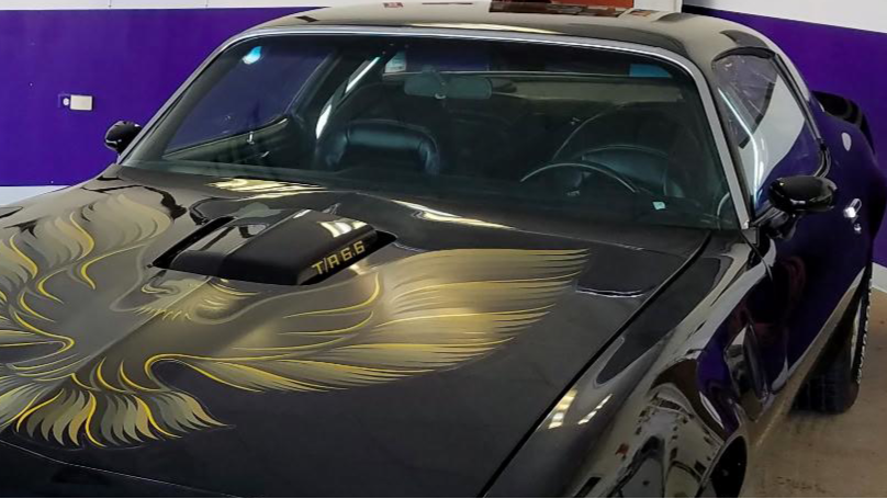 Black sports car with a large golden phoenix decal on the hood, inside a garage with purple walls.