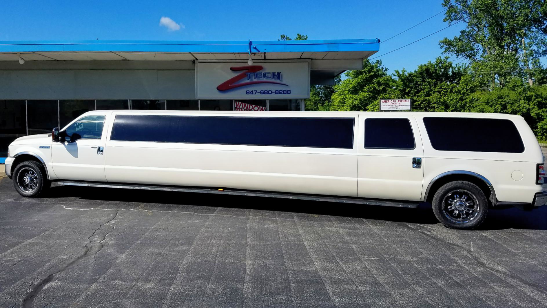 A white limousine parked in front of a business with a sign that reads 'Z Tech' and includes a phone number. The limousine is long with tinted windows and black wheels. Behind the vehicle are trees and a clear blue sky.
