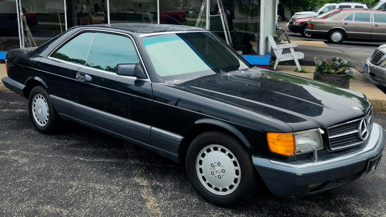 A black vintage Mercedes-Benz coupe parked outside a storefront with potted plants and other cars in the background.