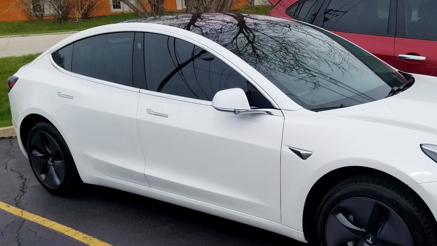 A white Tesla Model 3 parked in a parking lot, with a red vehicle behind it and a green grassy area with trees and buildings in the background.