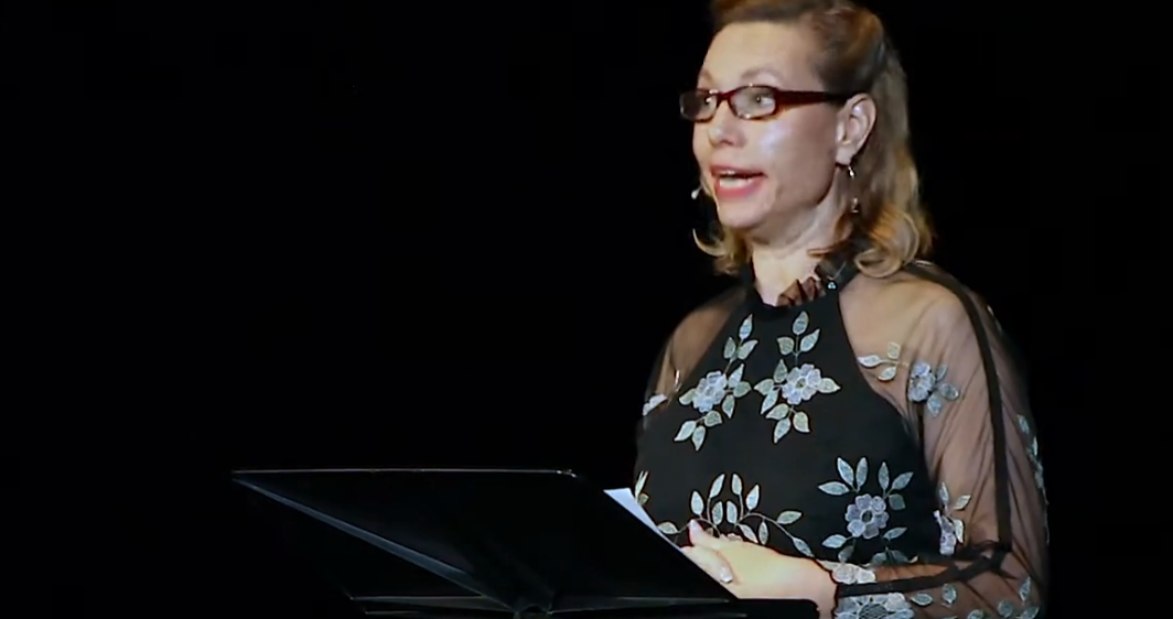 A woman wearing glasses and a black floral sheer top, speaking in front of a black background.