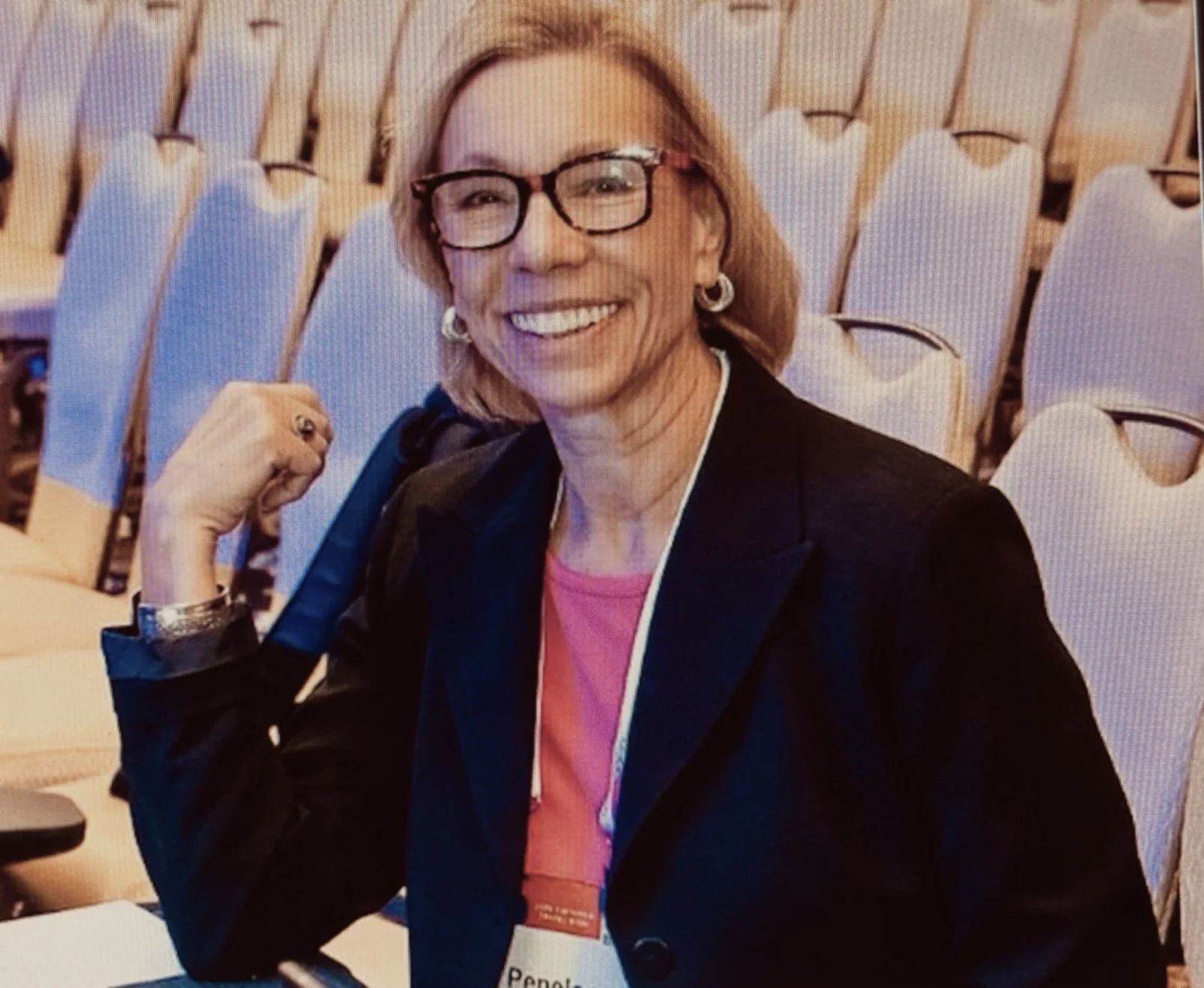 A smiling woman with blonde hair, glasses, and earrings, sitting in a conference room with empty chairs behind her. She is wearing a black blazer, pink shirt, and a wristwatch, and is making a fist with her right hand.