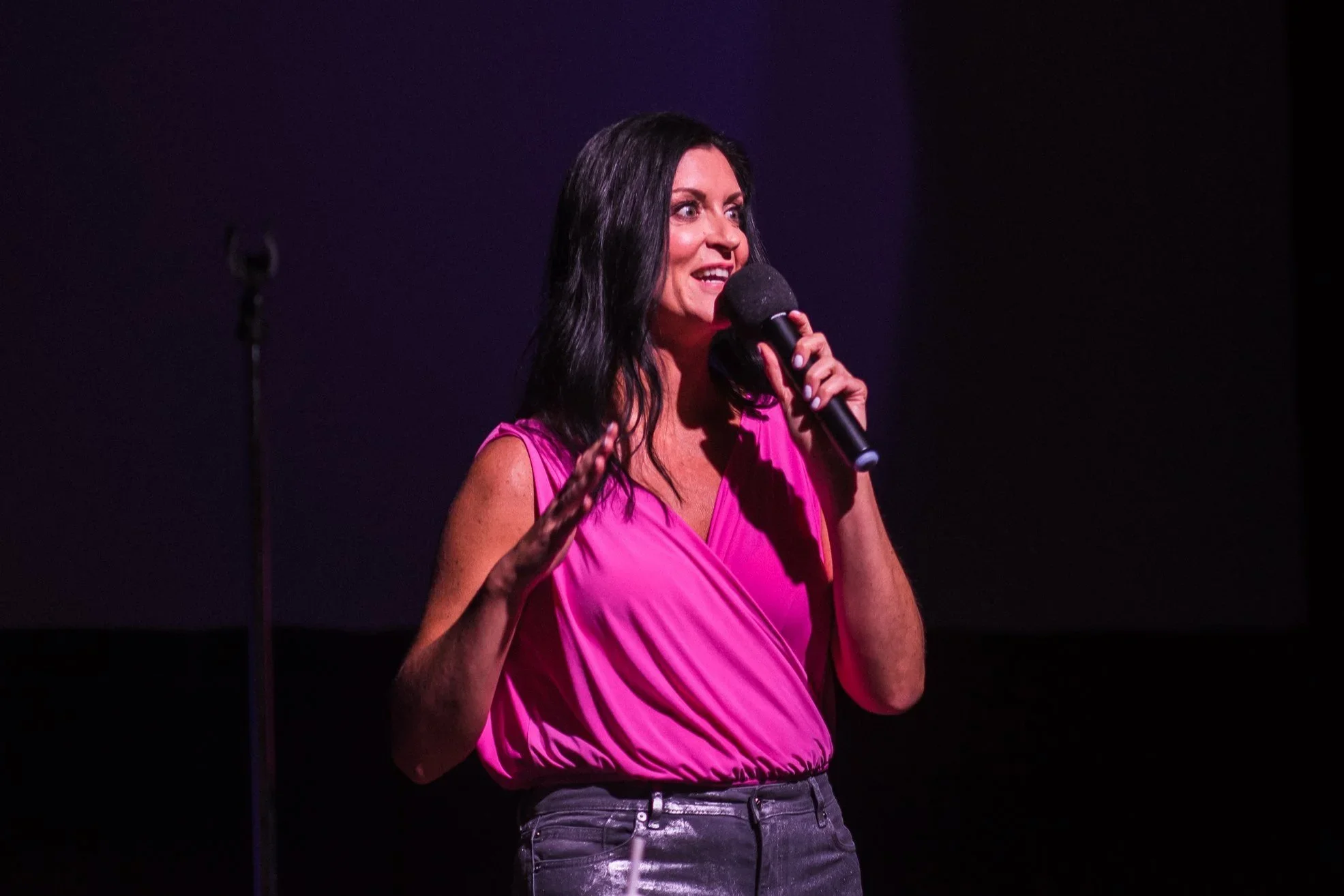A woman with dark hair wearing a bright pink sleeveless top is holding a microphone and speaking on stage. The background is dark.