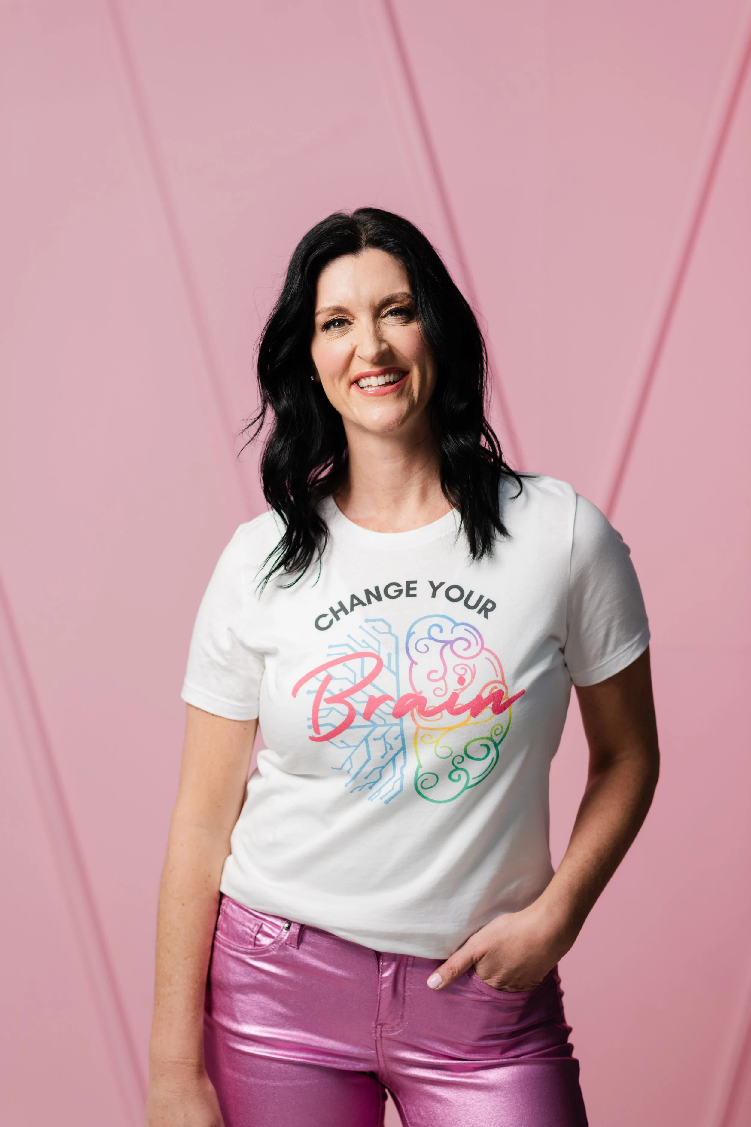 A woman with black hair, wearing a white T-shirt with colorful typography and designs, and metallic pink pants, standing in front of a pink geometric background.