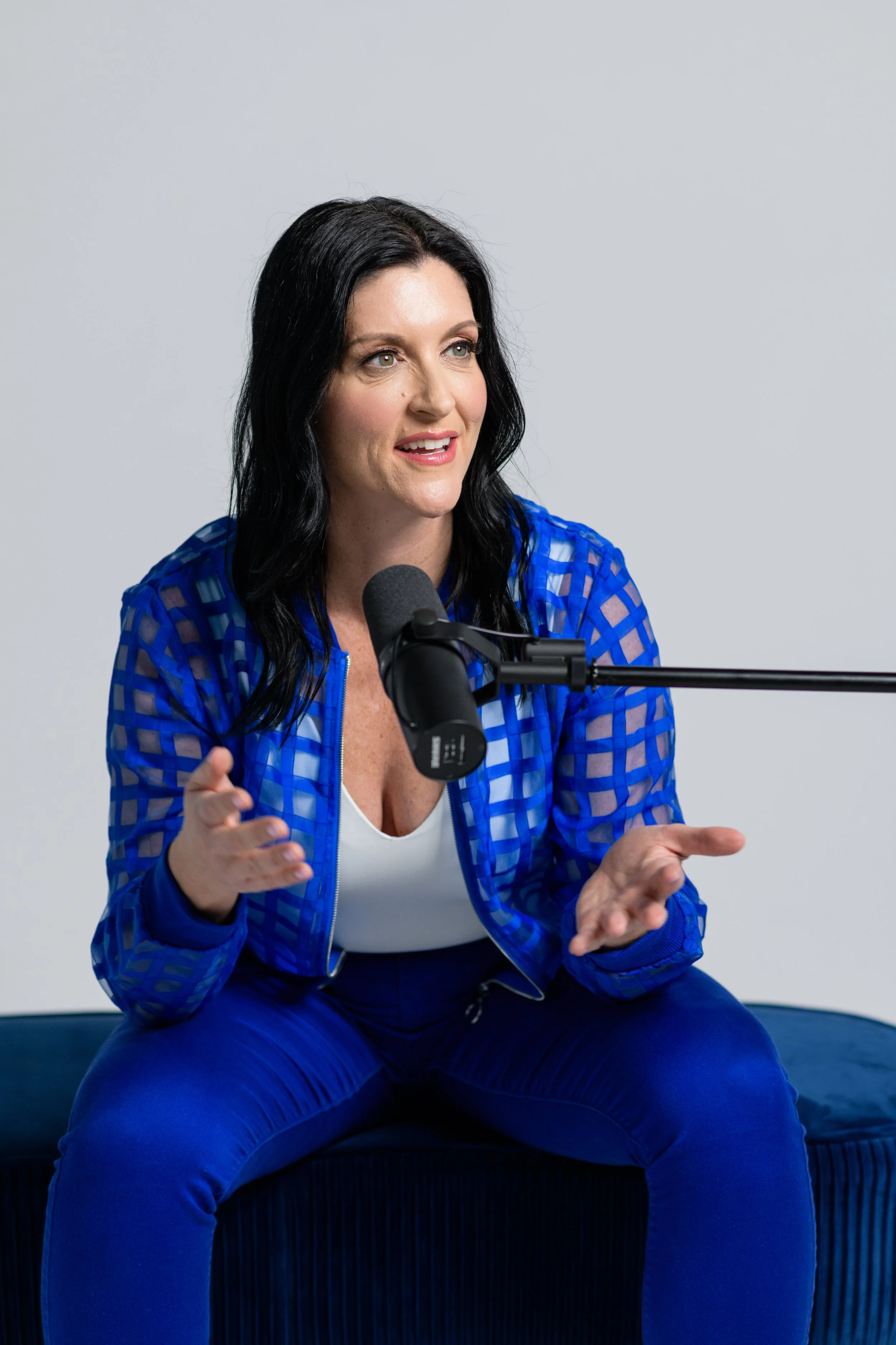 A woman with black hair, wearing a blue checkered jacket and blue pants, is sitting and speaking into a microphone, gesturing with her hands.
