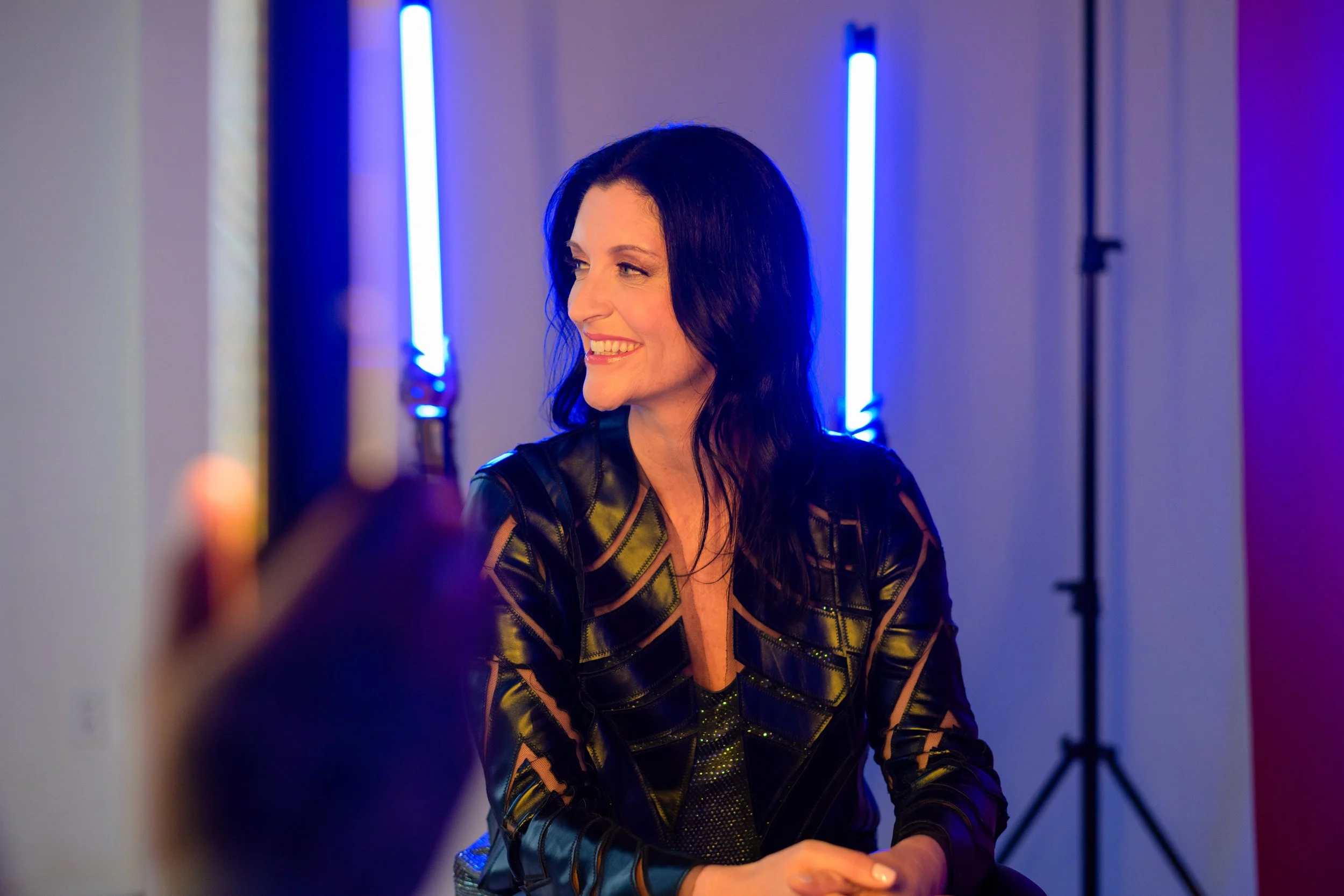 A woman with long dark hair smiling during a photoshoot, illuminated by blue and purple lighting.