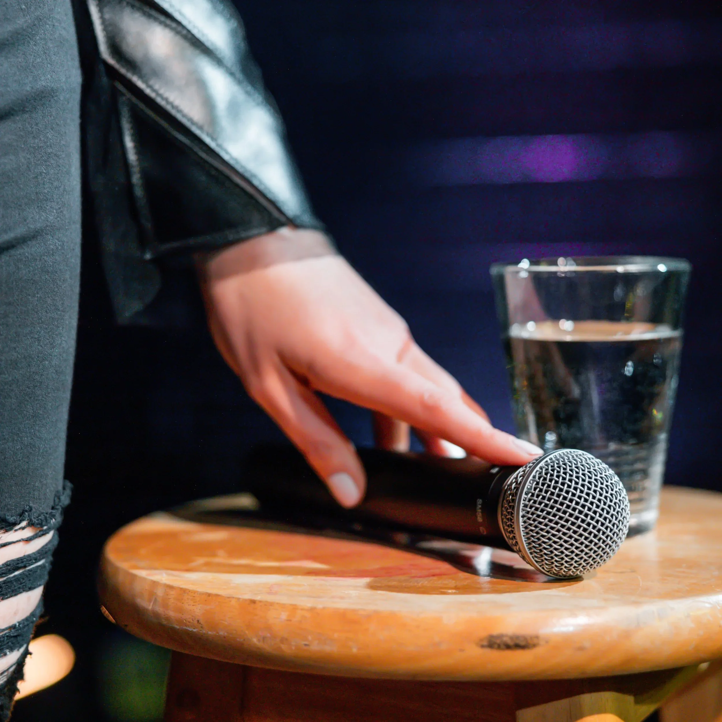 A person with a leather jacket sitting at a table, holding a microphone on a wooden surface, with a glass of water nearby.