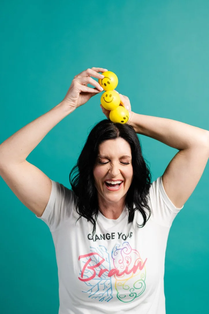 A woman, Jaime Armstrong, with black hair smiling with her eyes closed, holding a stack of yellow stress balls with smiling and surprised emojis above her head against a teal background.