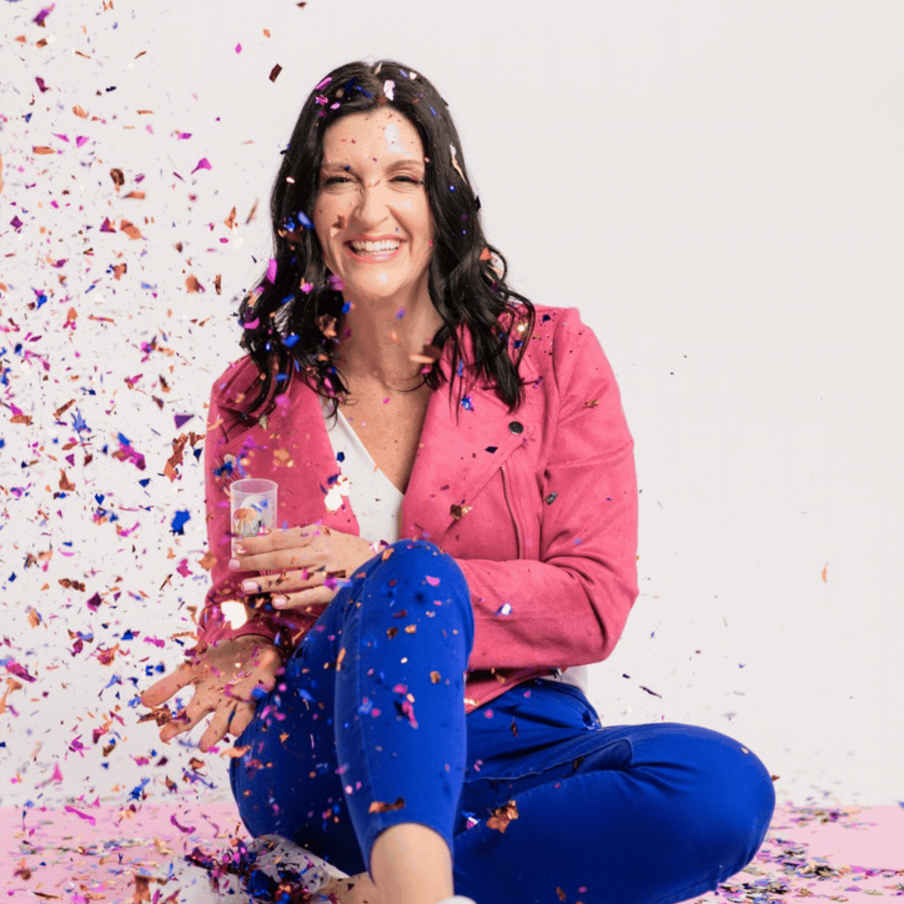 A woman, Jaime Armstrong, with dark hair wearing a pink jacket and blue pants is sitting on the floor, smiling, surrounded by colorful confetti, with some confetti still falling around her.